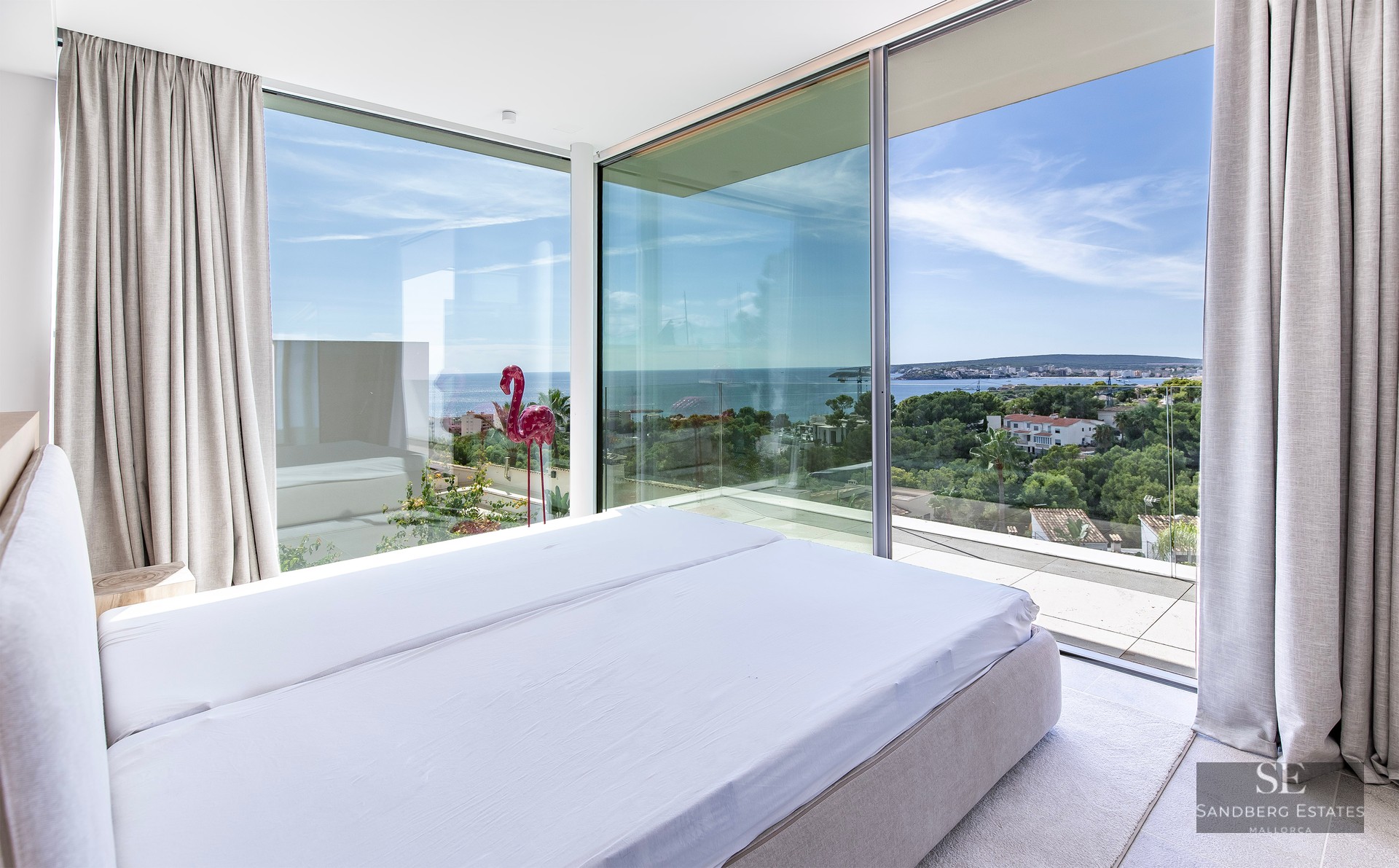 Modern bedroom with floor-to-ceiling corner windows overlooking the Mediterranean sea and coastal landscape.