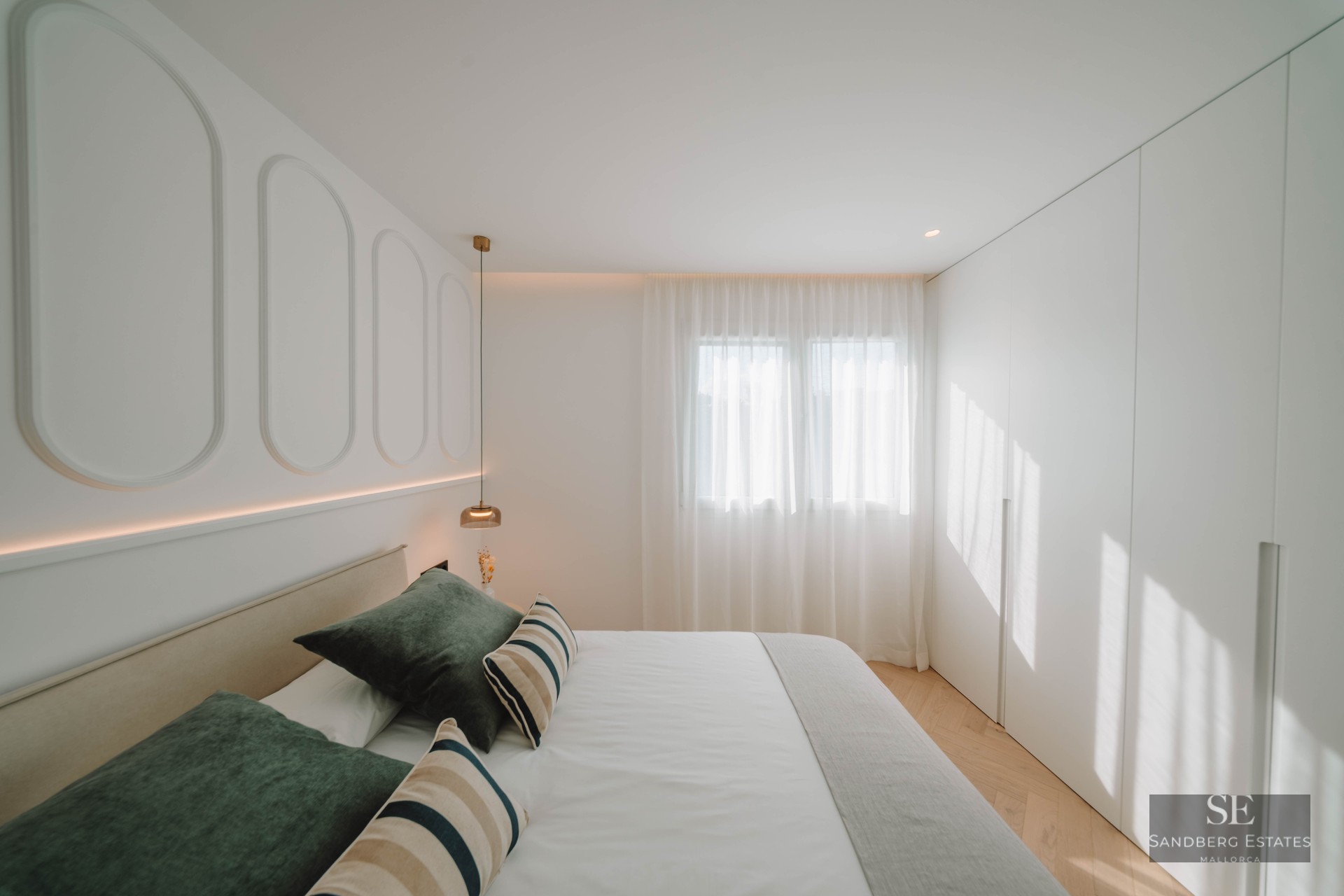 Bright bedroom with white paneled walls, a double bed with green pillows, herringbone wood floors, and built-in closets.