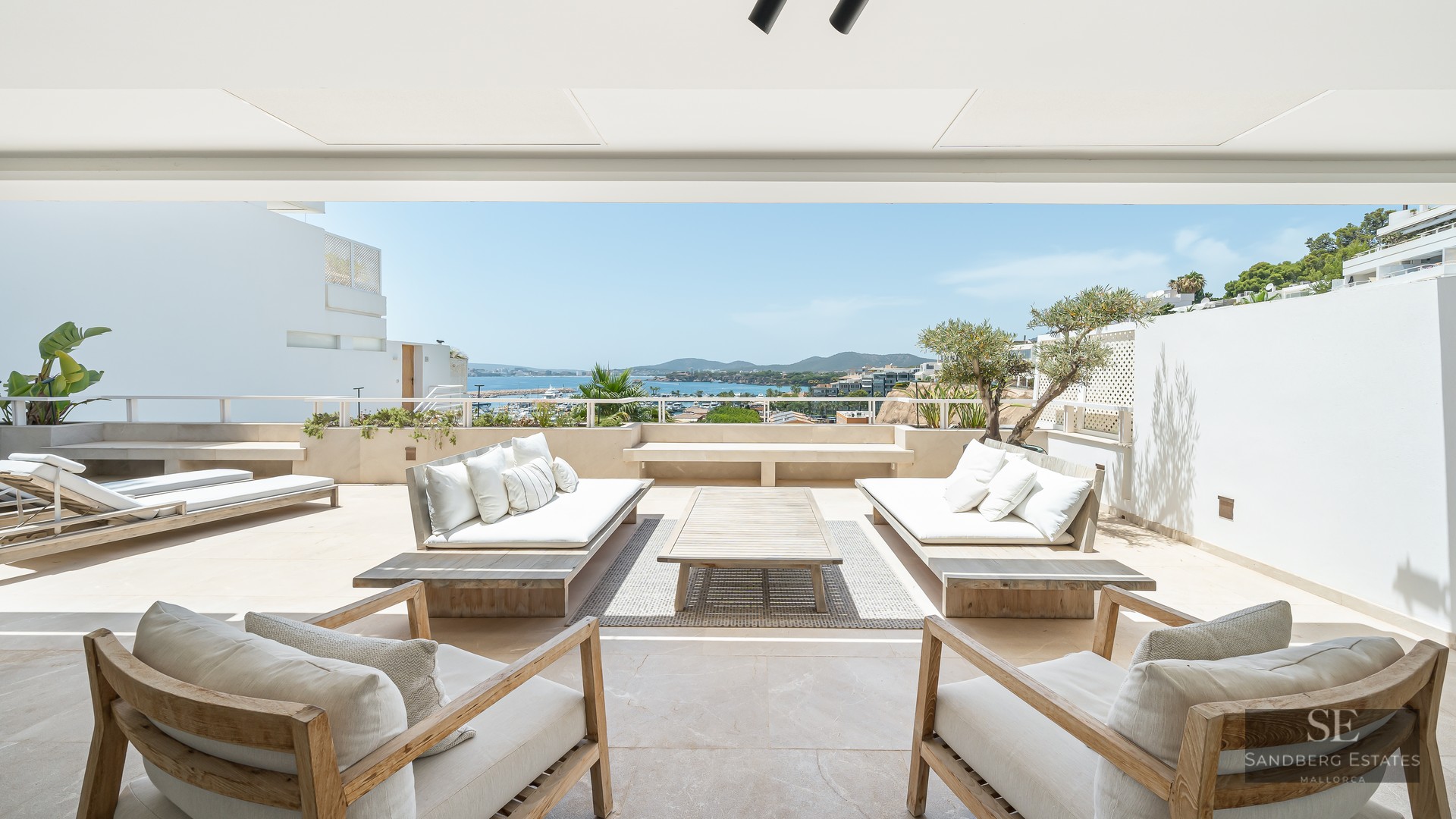 Spacious white terrace with wooden sofas and armchairs overlooking a blue sea and coastline under a bright sky.
