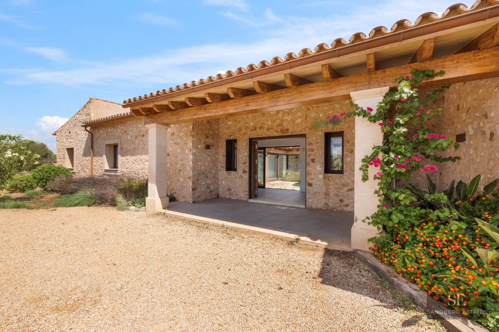 Entrance of a stone villa with a wooden porch, gravel path, and vibrant flowers under a blue sky.