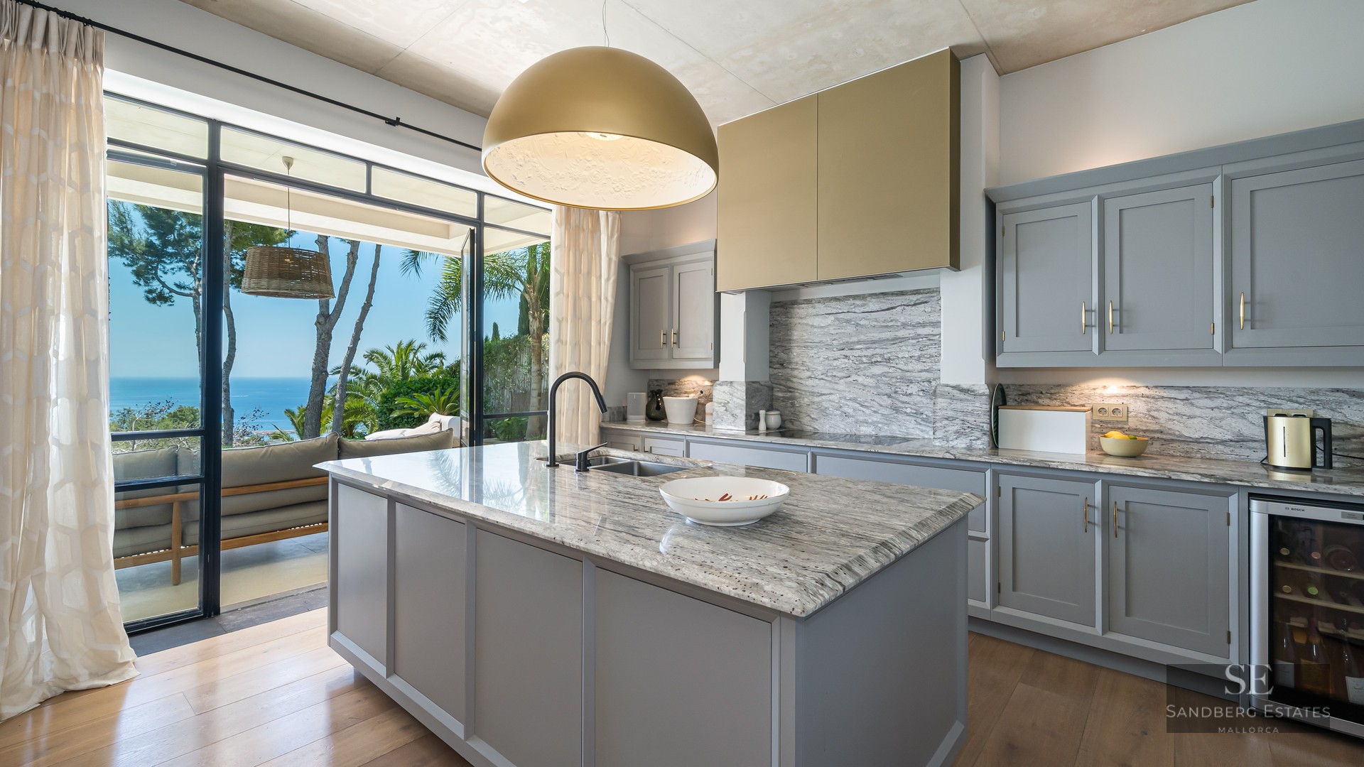 Modern kitchen with a marble island, grey cabinets, and a large open window overlooking the Mediterranean sea.