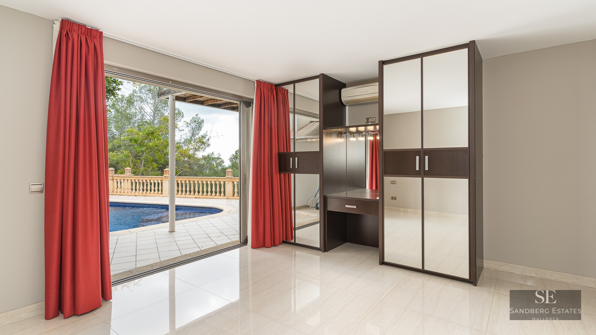 Modern room with red curtains, mirrored wardrobes, and a vanity, overlooking a swimming pool through glass doors.
