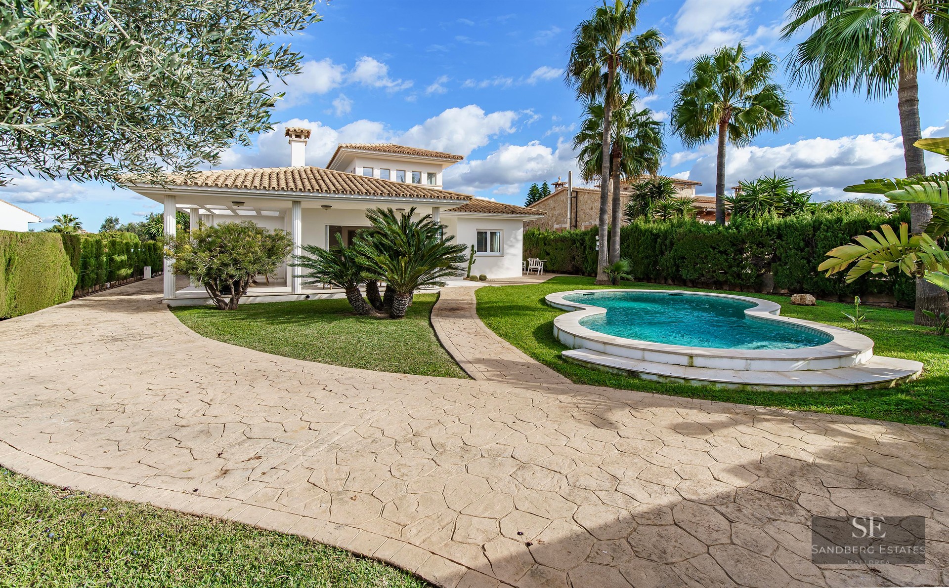 A Mediterranean villa with a curved private pool, lush green lawn, palm trees, and a stone pathway under a blue sky.