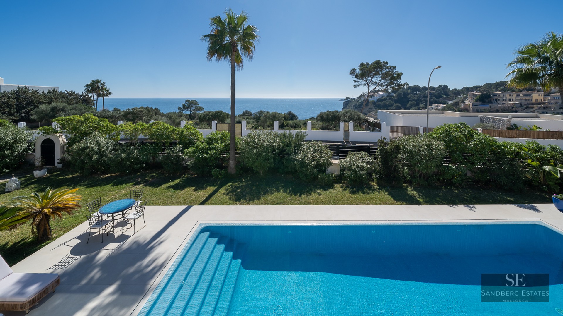 A blue tiled swimming pool and garden area overlooking the Mediterranean Sea under a clear blue sky.