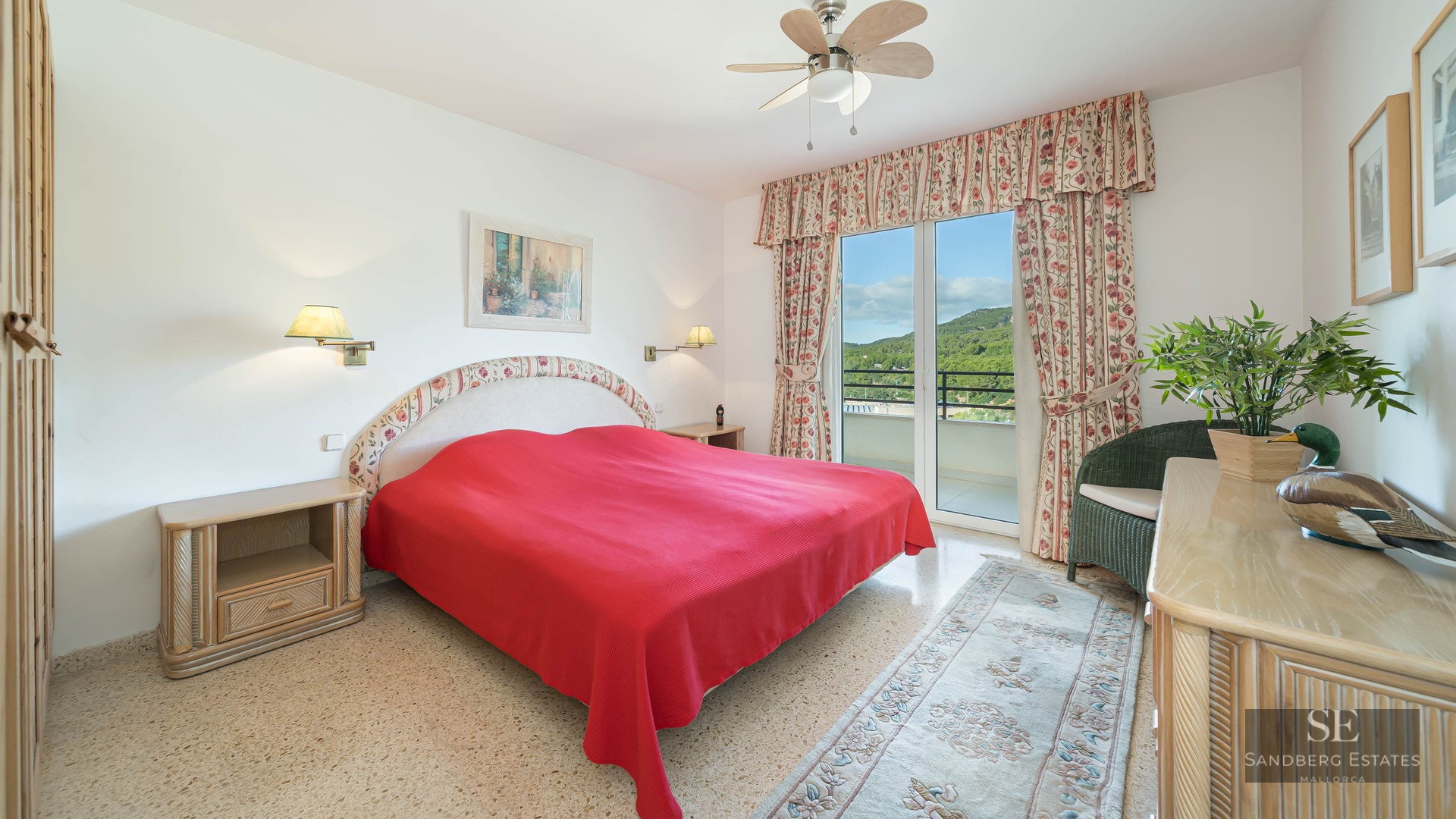 Bright bedroom featuring a red bedspread, floral curtains, and glass doors opening to a balcony with green hill views.
