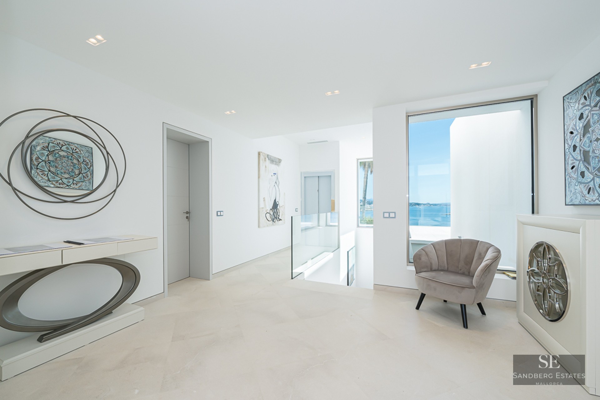 Bright modern hallway with stone floors, glass railings, an elevator, and a large window overlooking the blue sea.