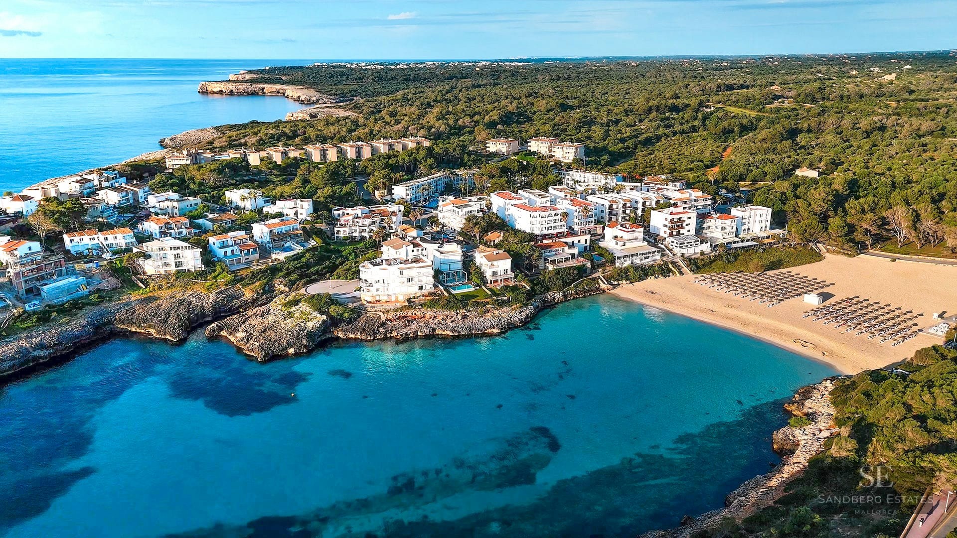 Aerial view of a turquoise bay with a sandy beach and white buildings nestled between rocky cliffs and green forest.