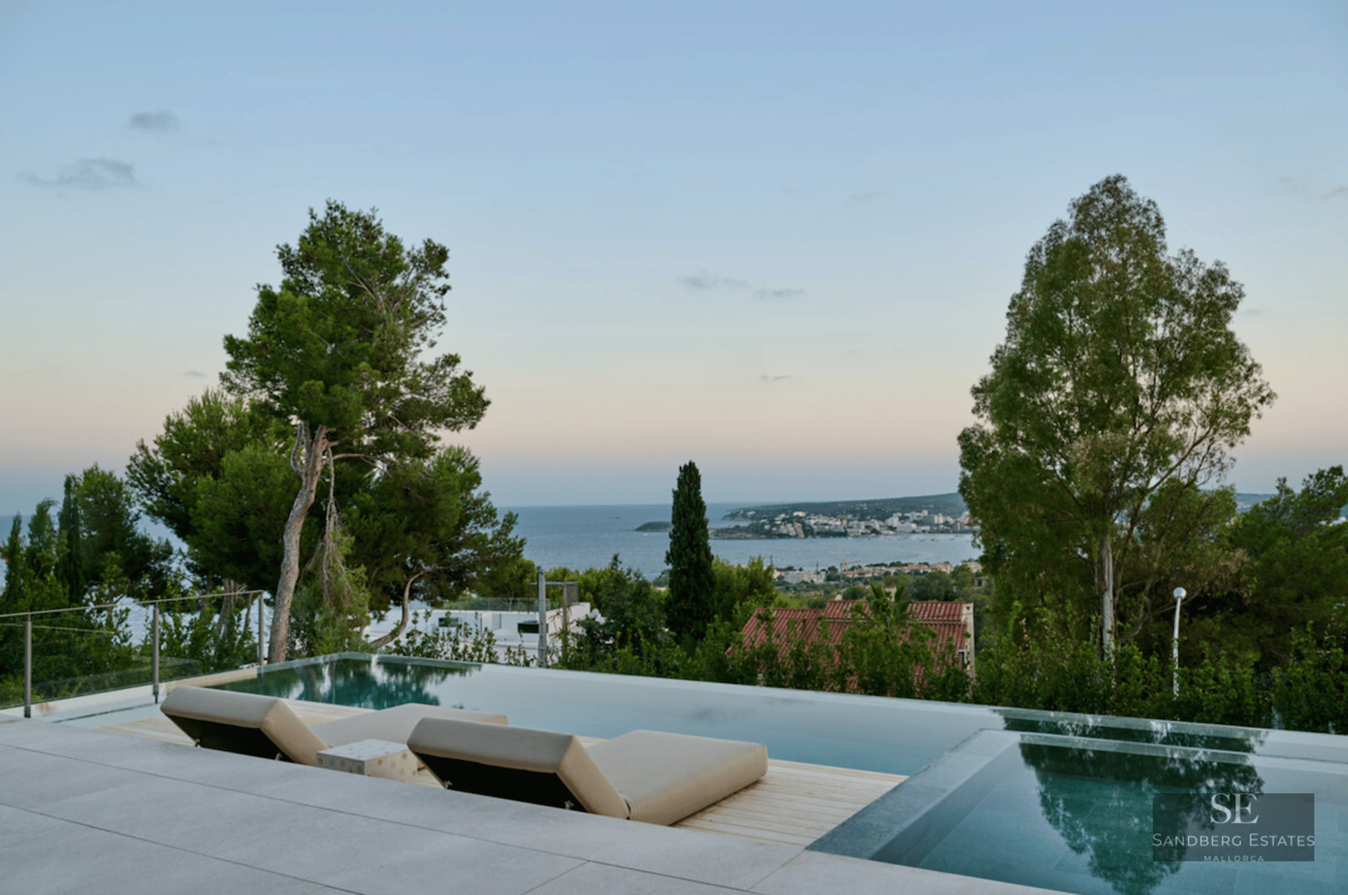Modern infinity pool with two beige loungers overlooking a calm blue sea and coastal town at dusk.
