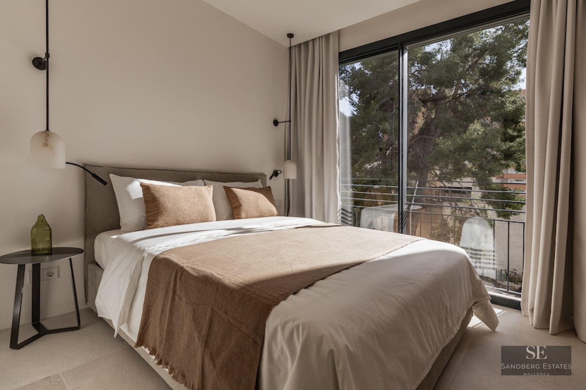 Bright bedroom with a large bed, neutral linens, designer pendant lights, and a floor-to-ceiling window viewing trees.