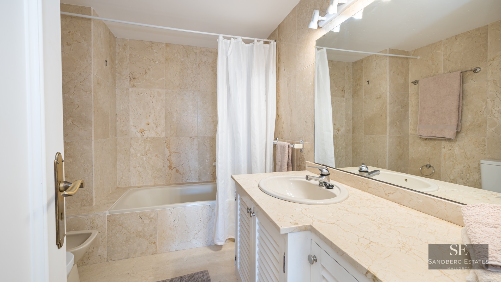 Bathroom featuring beige marble walls, a large mirror, white vanity with marble top, and a bathtub.