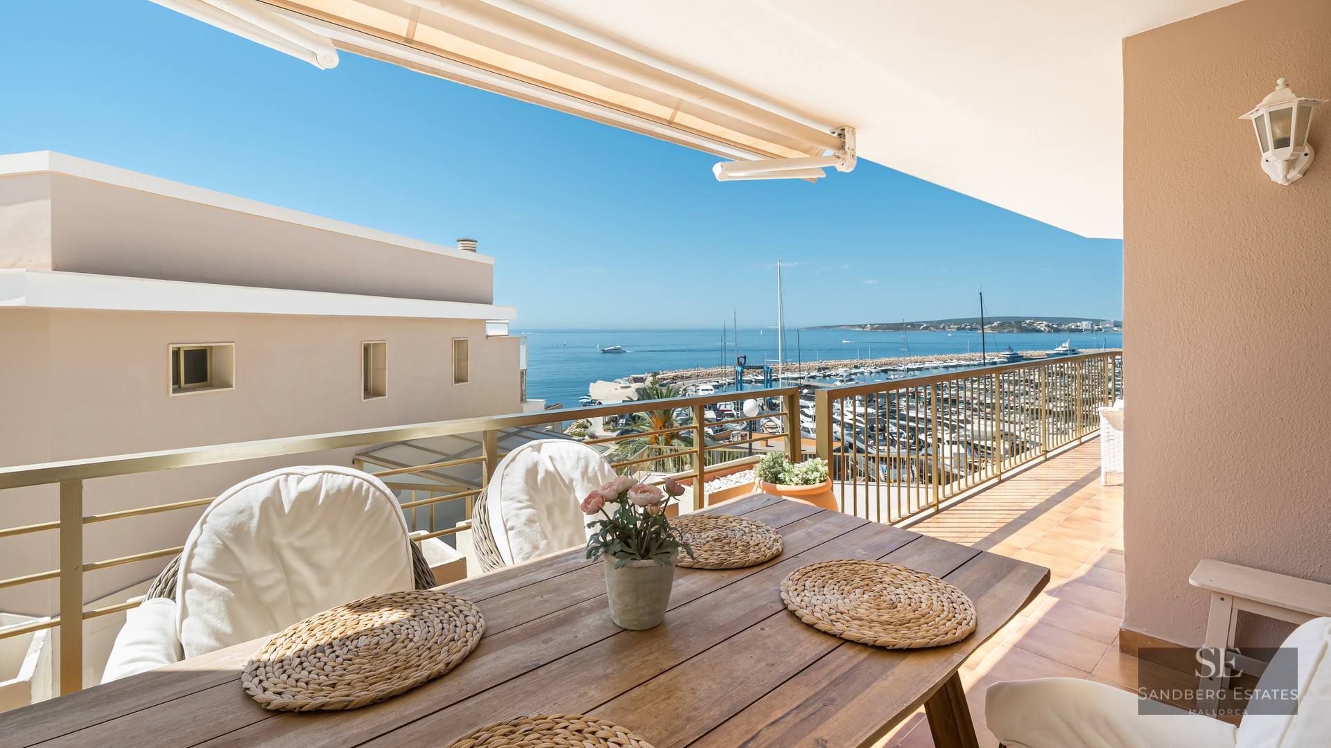 Balcony with wooden dining table and white chairs overlooking a marina and blue sea under a clear sky.