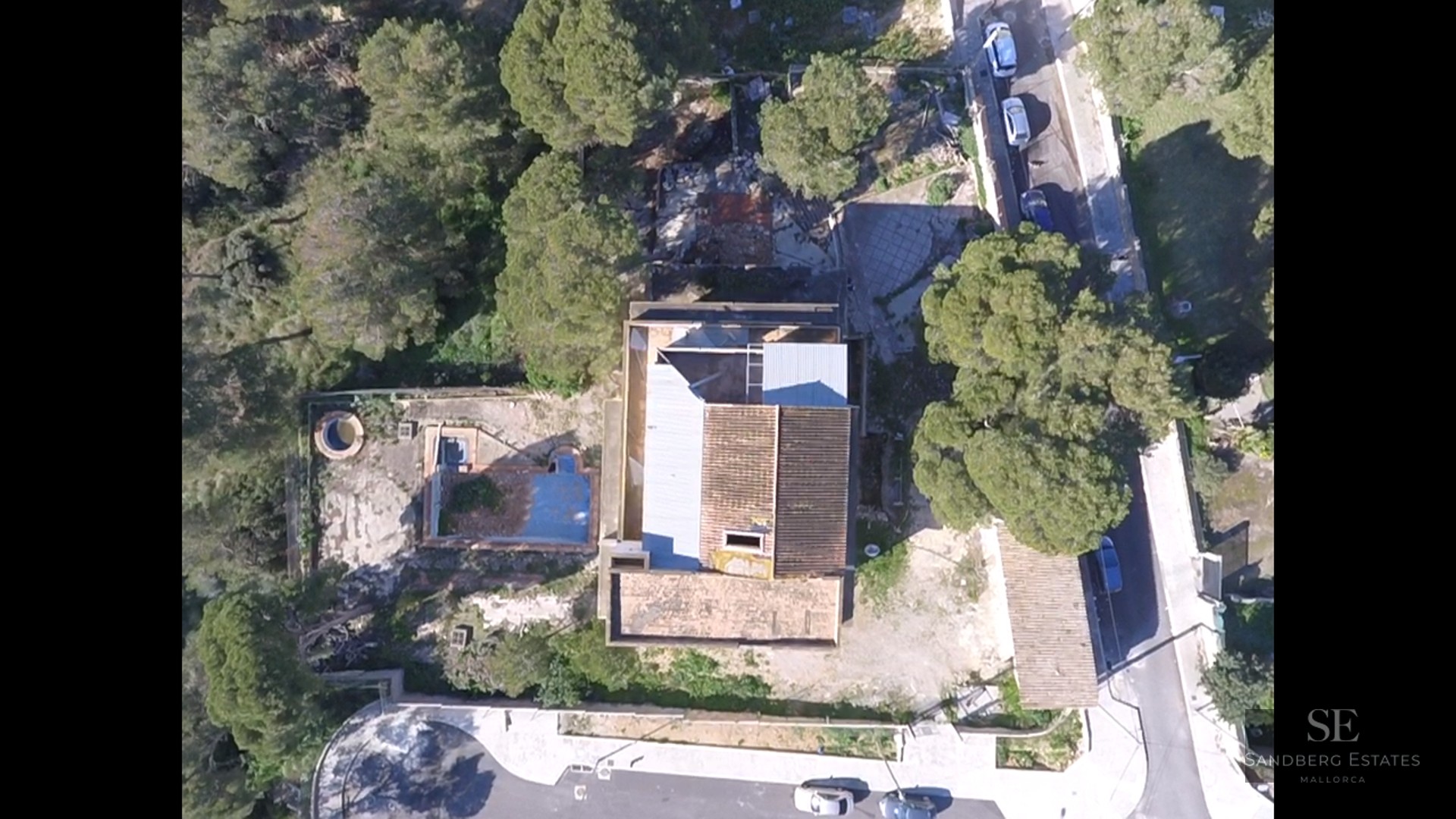 Top-down aerial view of a house with tiled roof, empty swimming pool, and surrounding pine trees.