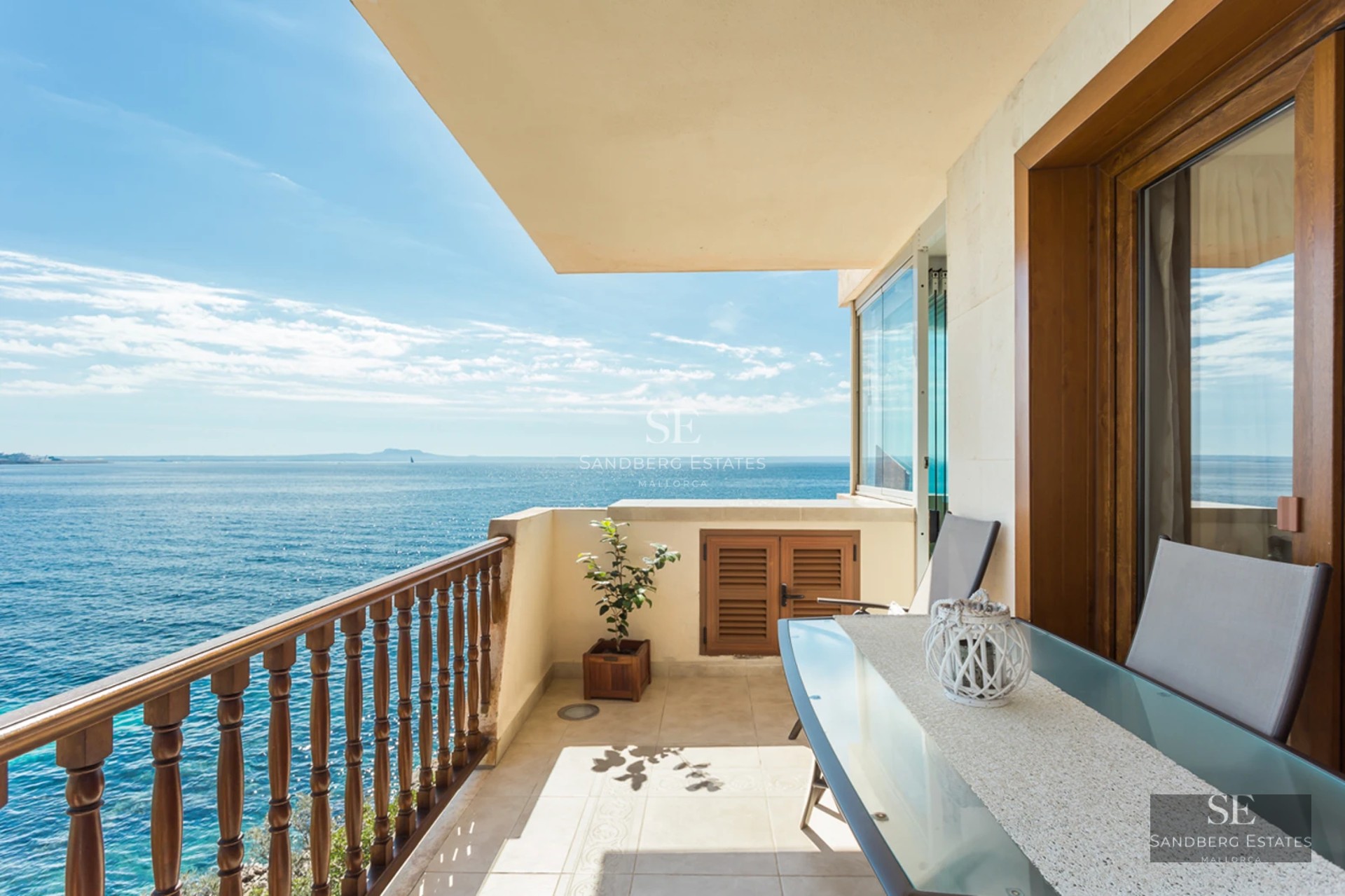 A sunny balcony with a glass dining table and wooden railing overlooking a vast blue ocean under a clear sky.