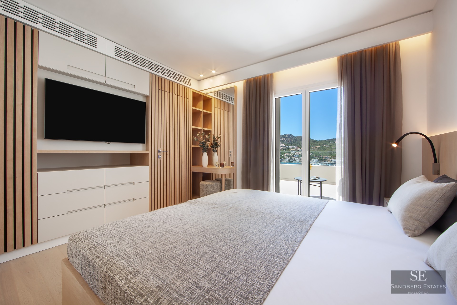 Bright bedroom with large bed, wood slat panels, wall-mounted TV, and balcony overlooking coast and mountains.
