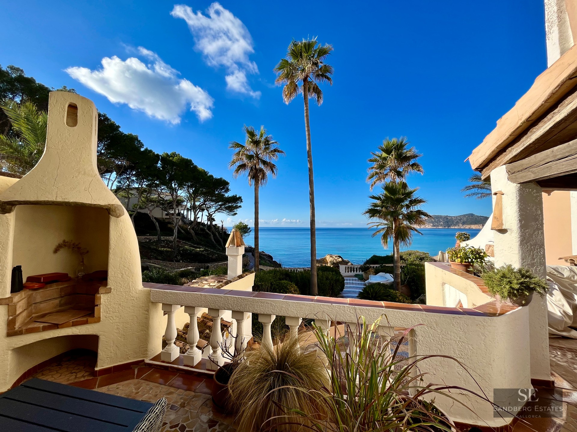 Terrace with a white built-in outdoor fireplace and balustrade overlooking palm trees and the vibrant blue sea.