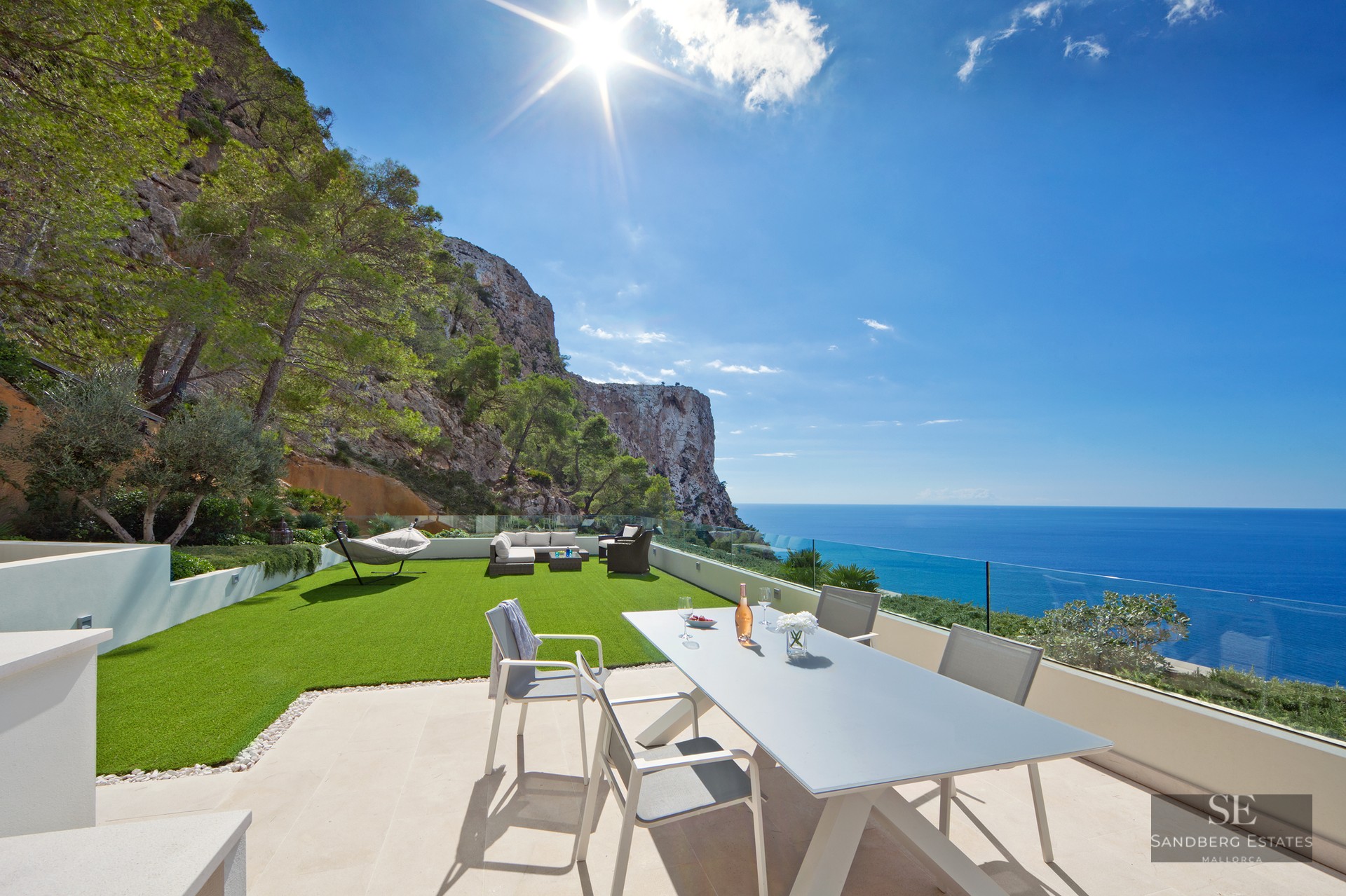 Modern outdoor terrace with dining area, artificial lawn, and glass railing overlooking the sea and cliffs.