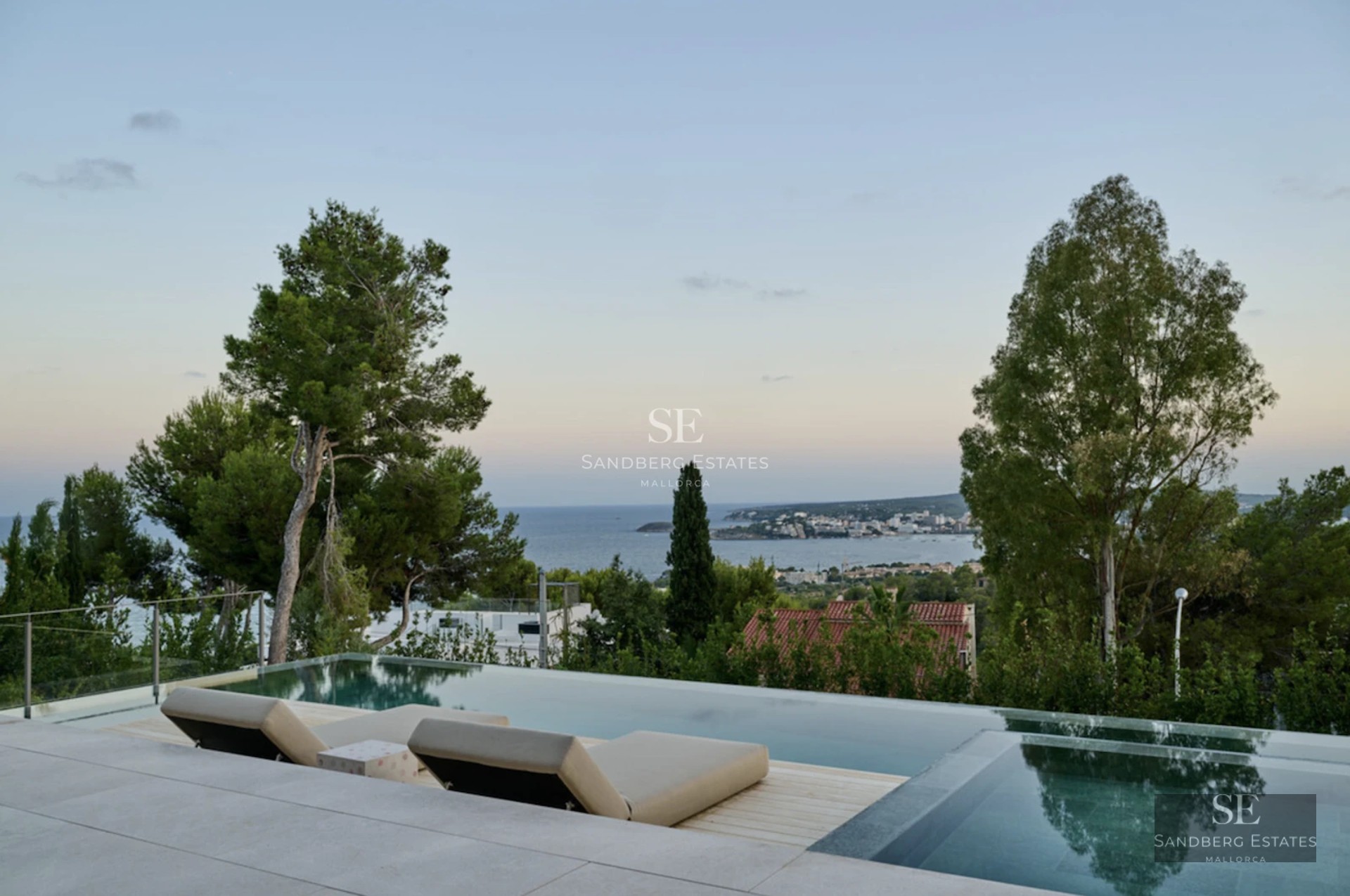 View of an infinity pool surrounded by lush greenery, with a modern house in the background and clear blue sky.