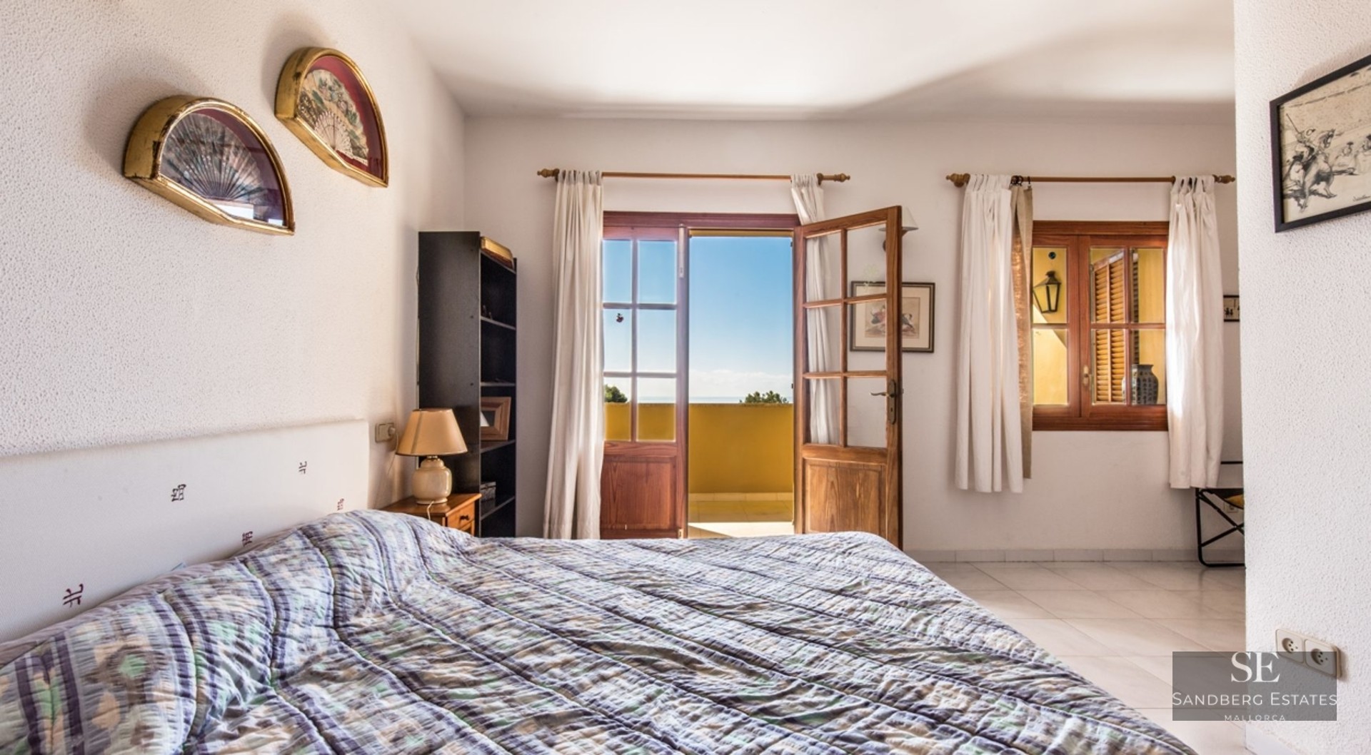 A bright bedroom with a double bed, white textured walls, and open wooden doors leading to a sunny balcony with a view.