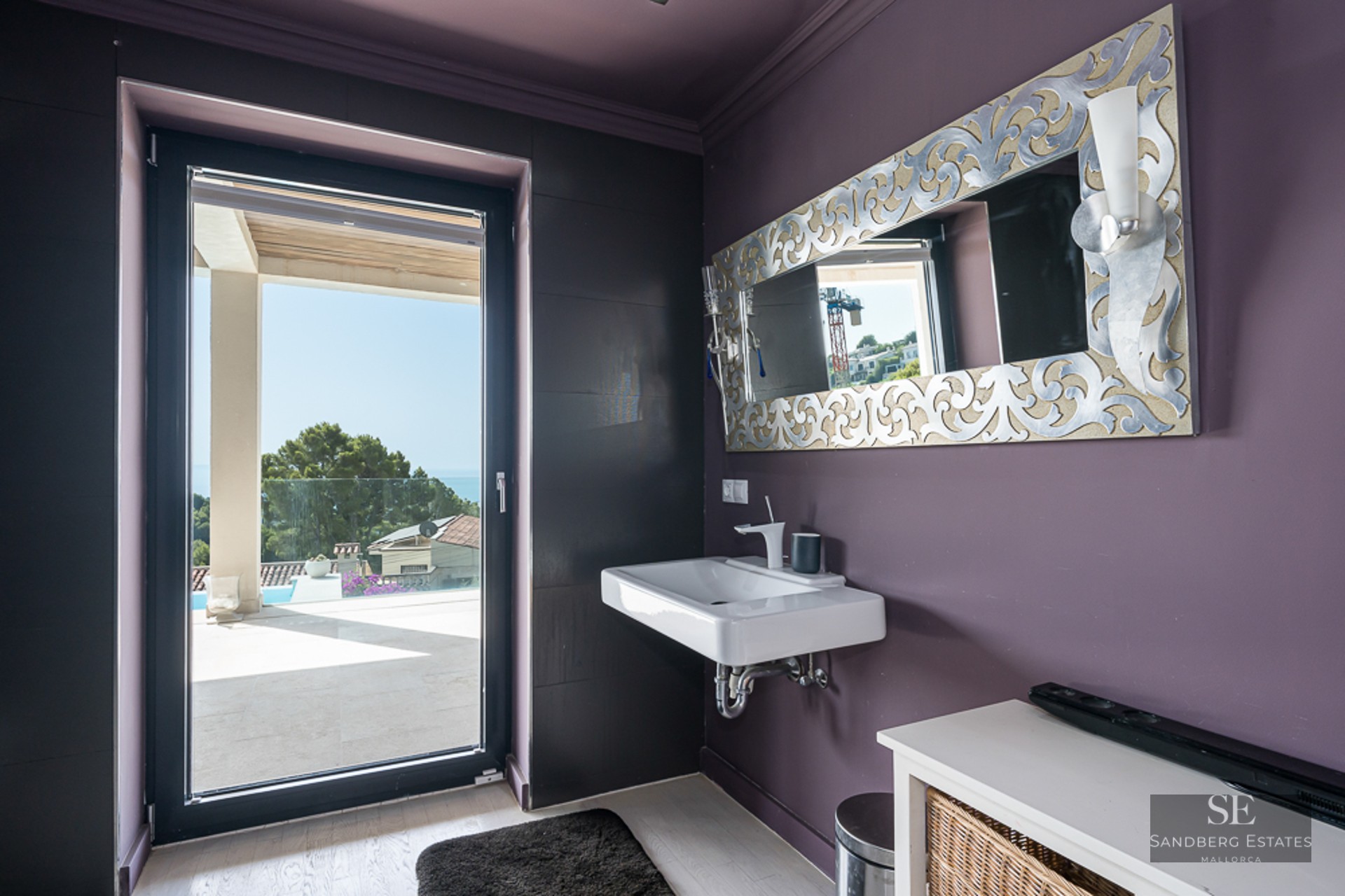 Modern bathroom with plum-colored walls, floating white sink, ornate silver mirror, and glass door to a sunny terrace.