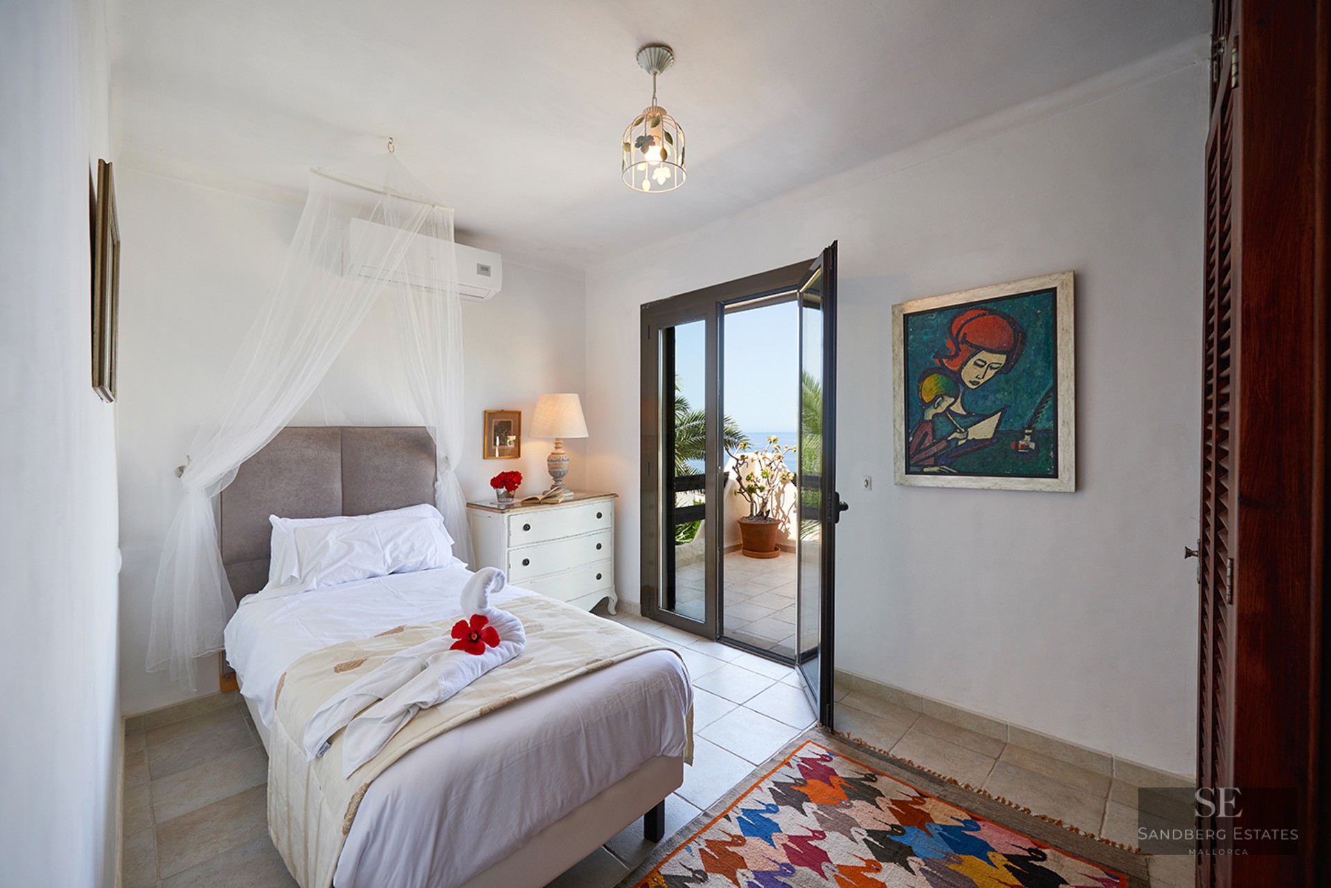 A white bedroom with a mosquito net over the bed, a colorful rug, and open glass doors leading to a sunny terrace.