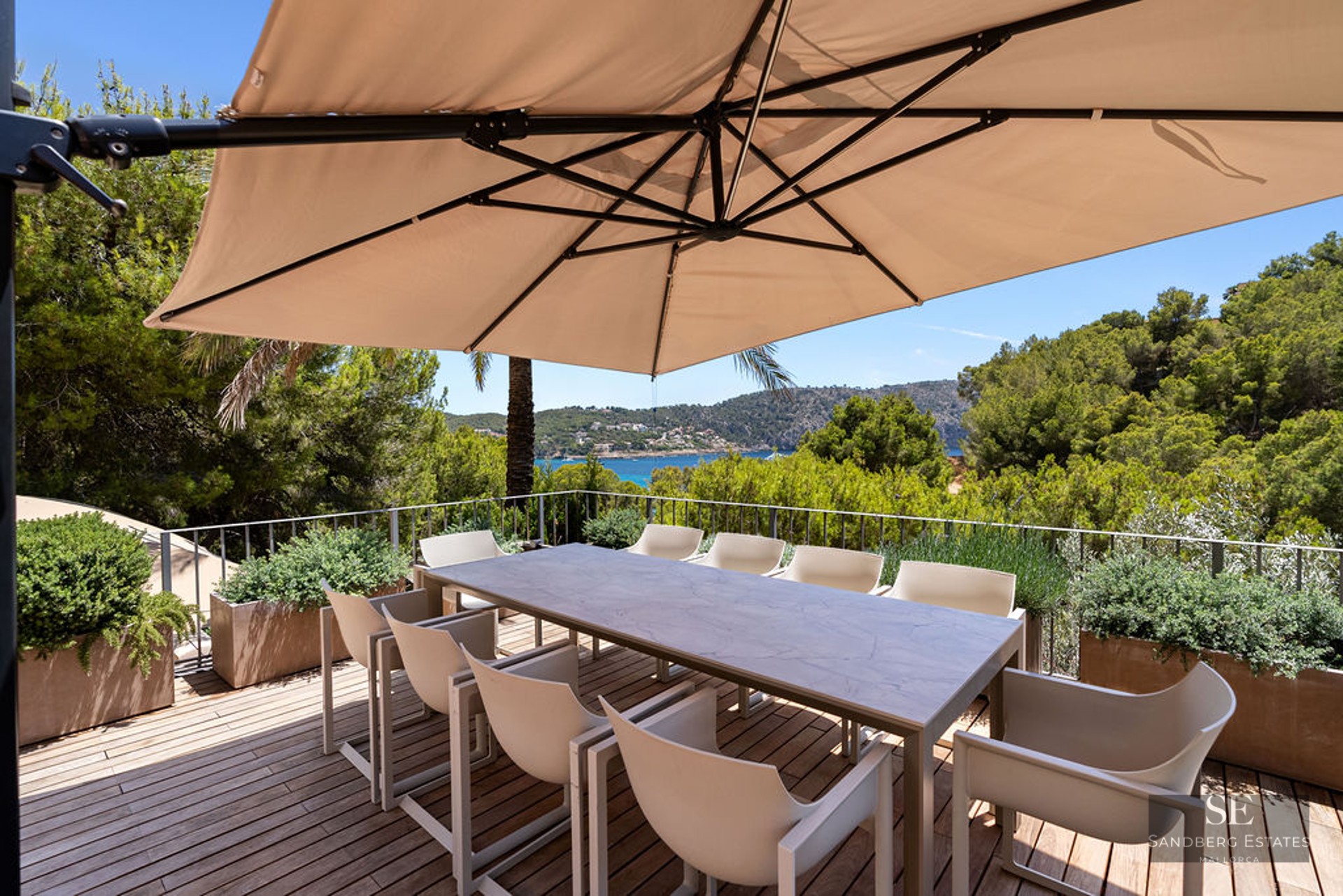Outdoor wooden terrace with a dining table and chairs under a large umbrella overlooking a sea bay.