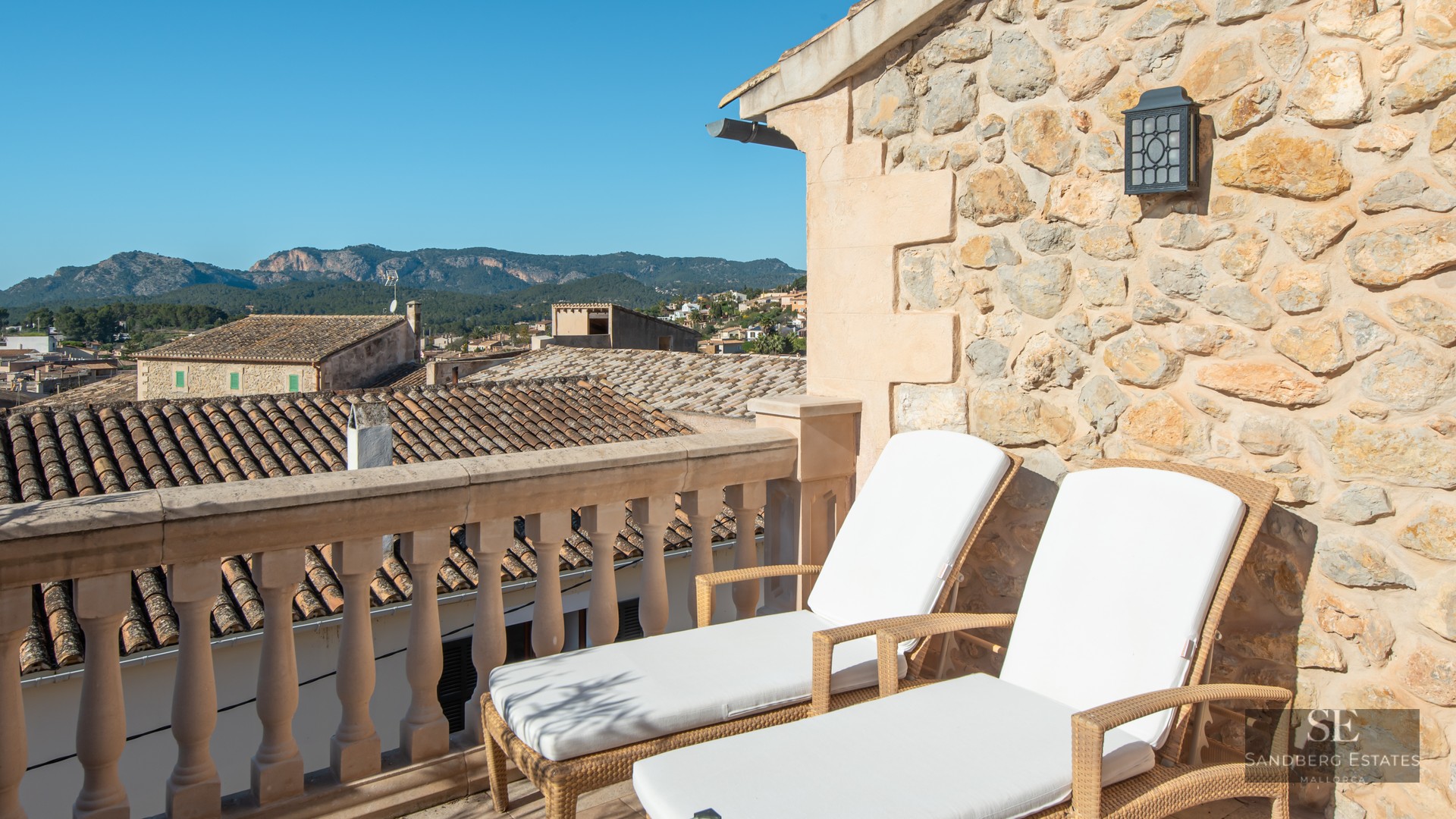 Two white sun loungers on a stone terrace overlooking a Mediterranean village and mountains.