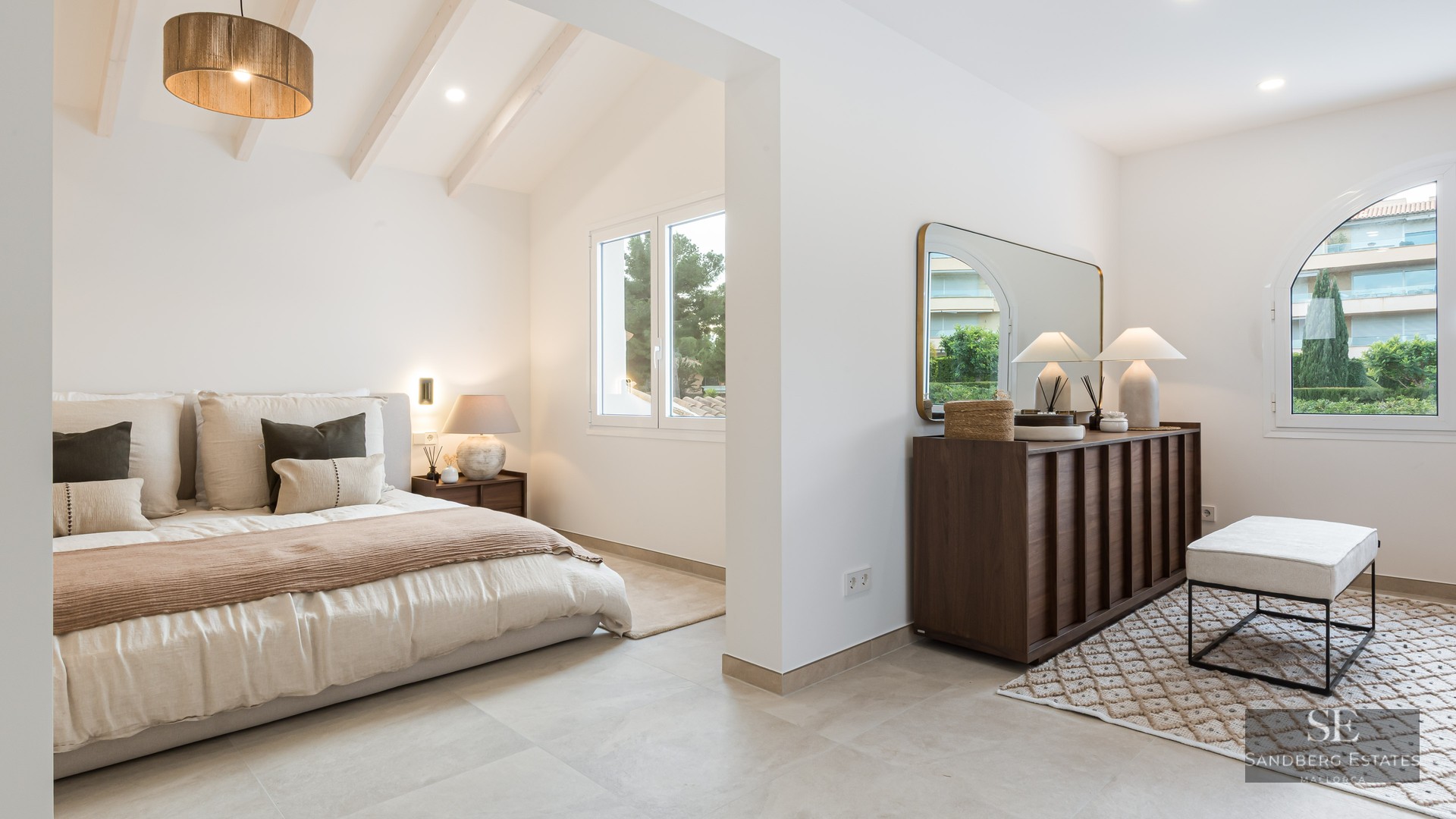 Modern master bedroom with white beamed ceilings, king bed, dark wood dresser, and natural light from large windows.