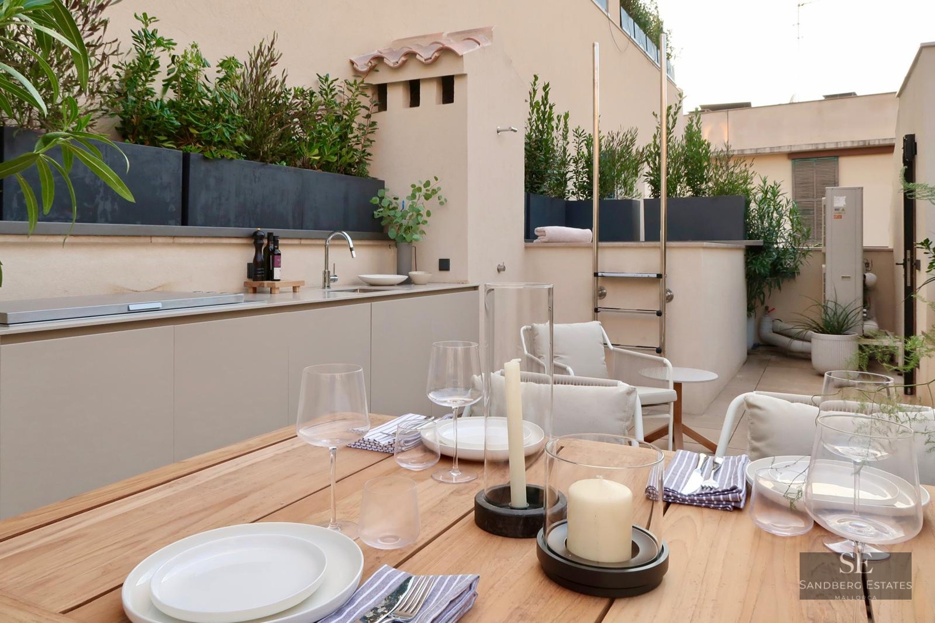 Wooden dining table on a modern terrace featuring an outdoor kitchen and lush greenery.