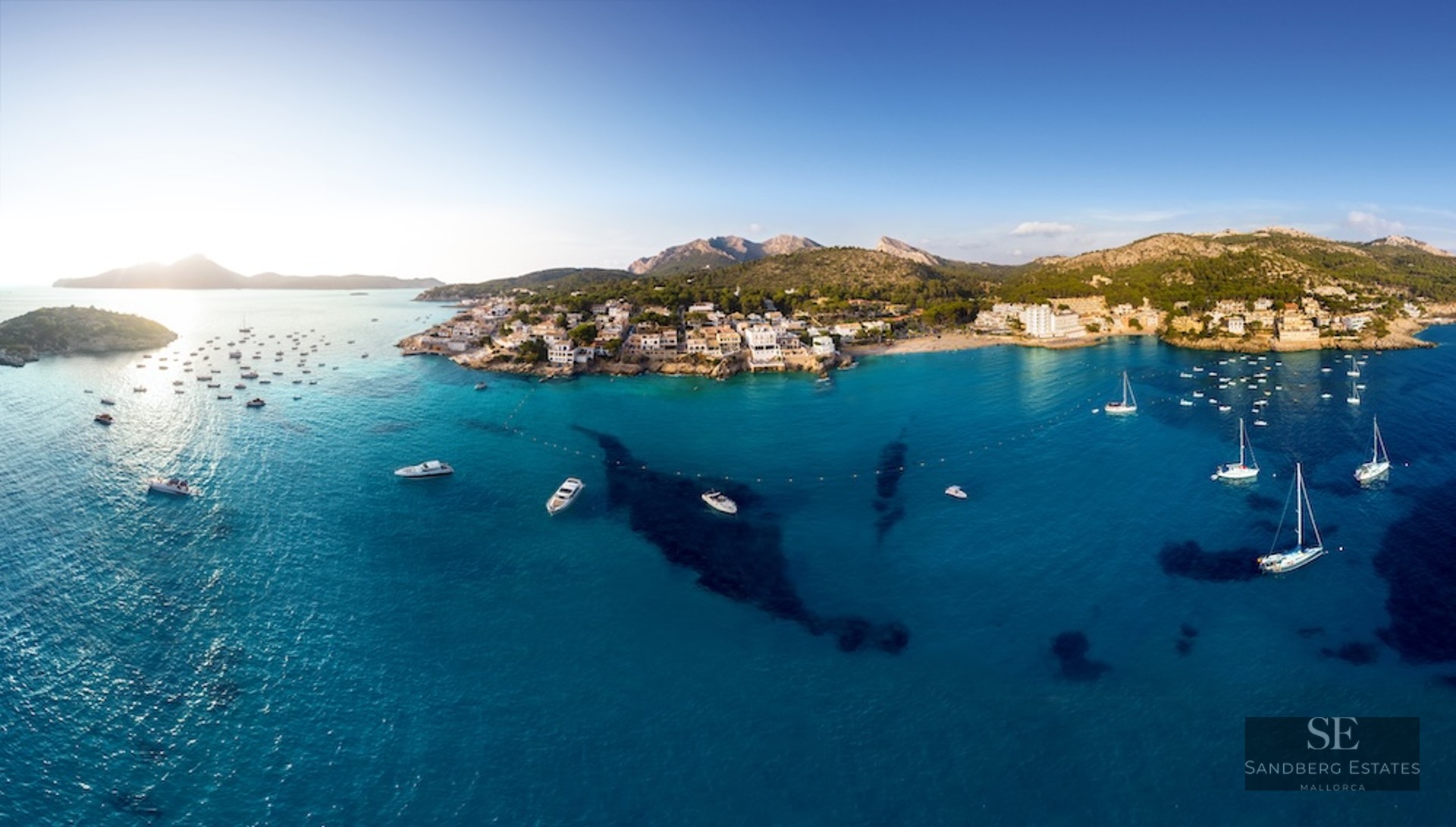 Aerial panorama of a Mediterranean bay with turquoise water, white boats, and a coastal town against green mountains.