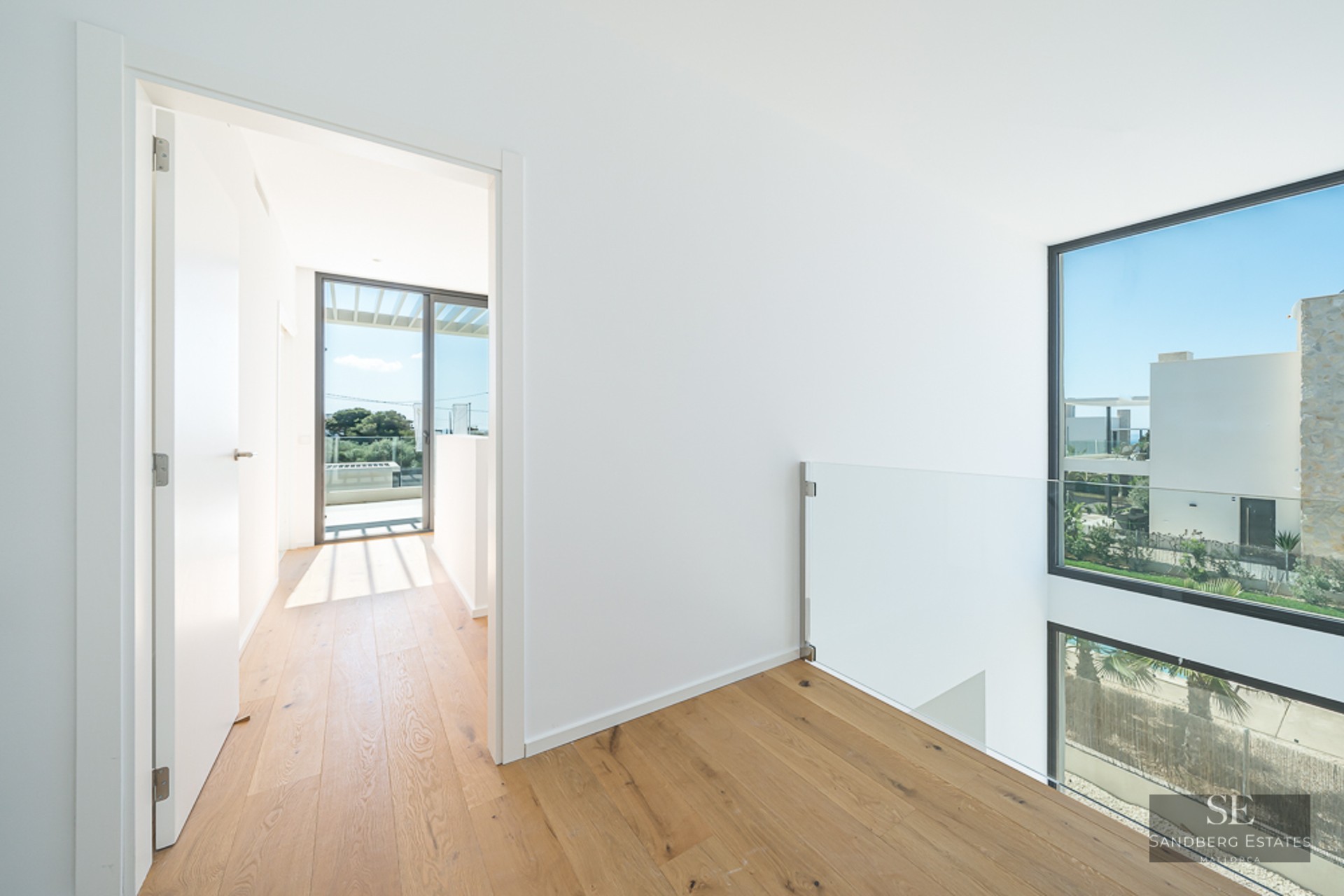 Bright hallway with light wood flooring, glass railing, and views through large windows to a sunny exterior.