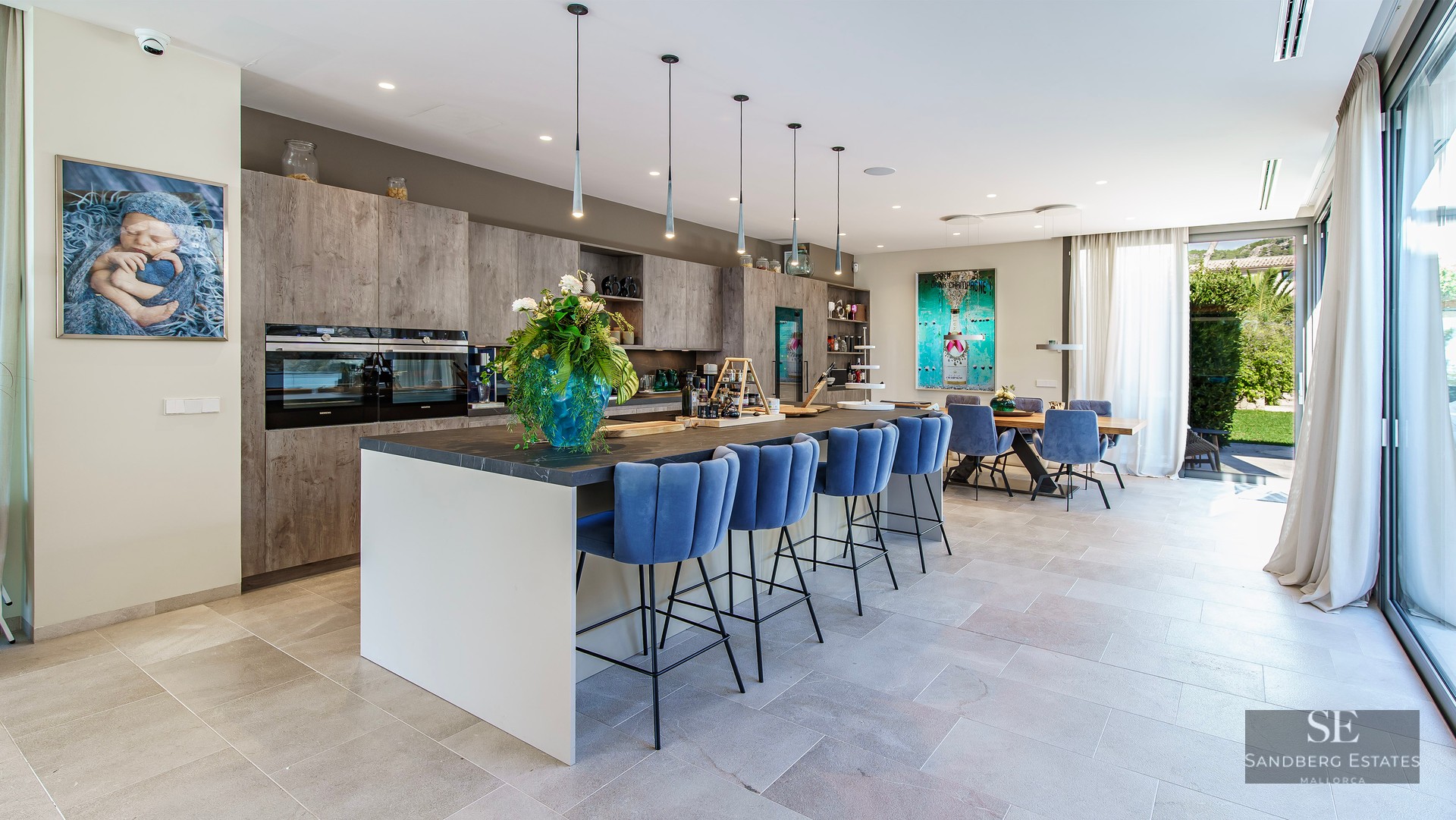 Cuisine moderne avec îlot central, tabourets en velours bleu et salle à manger intégrée baignée de lumière.