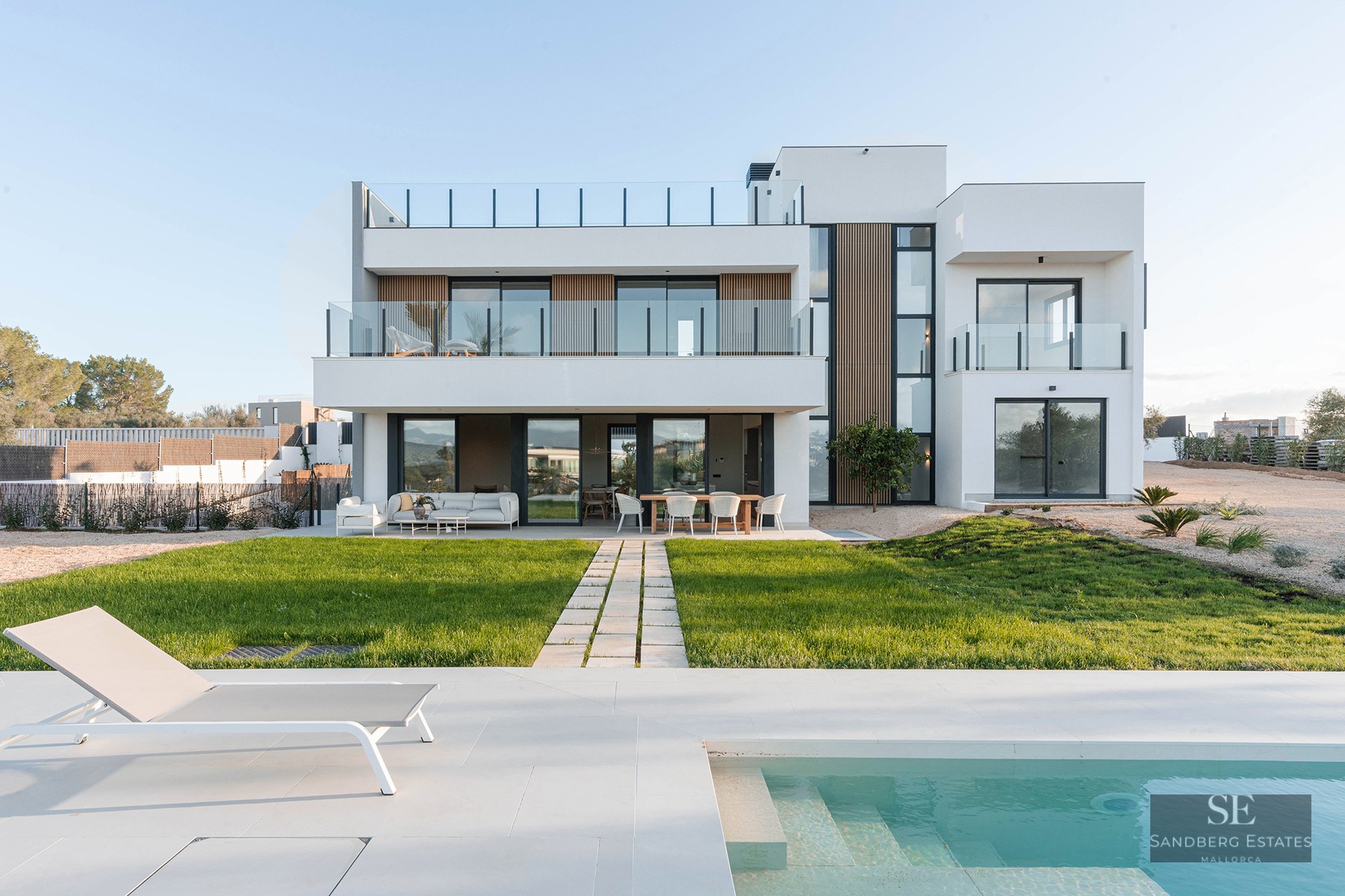 Modern white two-story villa with a turquoise swimming pool, sun lounger, and a green lawn under a clear sky.