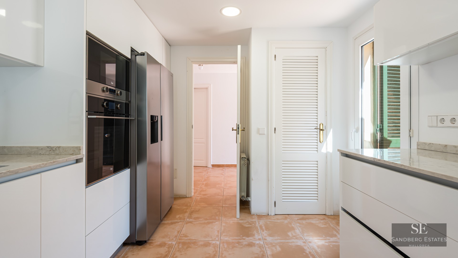 Bright kitchen with white cabinets, marble countertops, stainless steel fridge, and terracotta tile flooring.