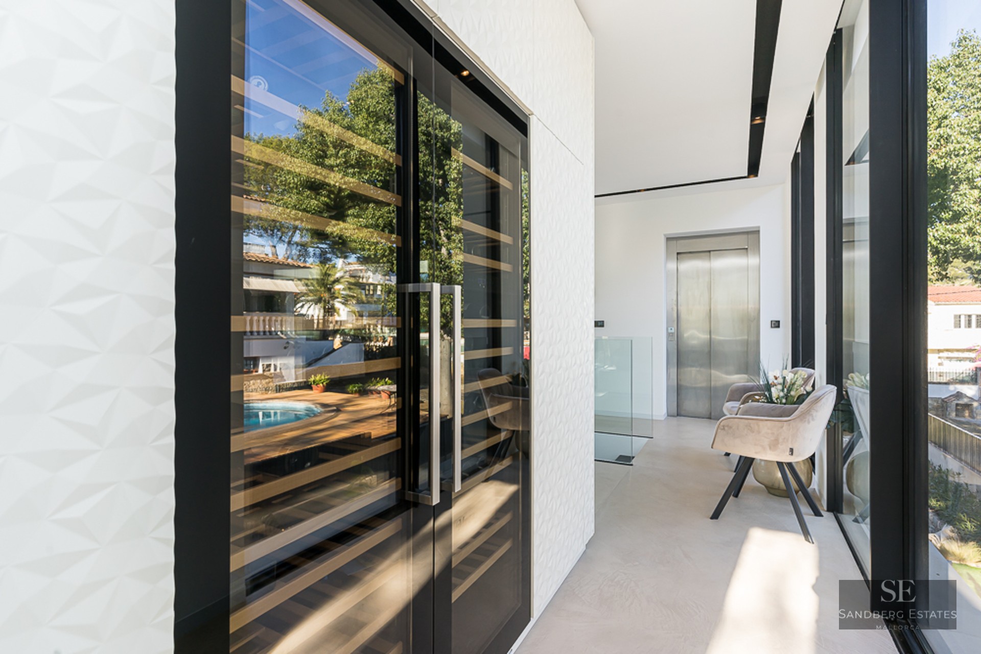Modern built-in wine cellar with glass doors reflecting a pool, next to a textured wall and an elevator.