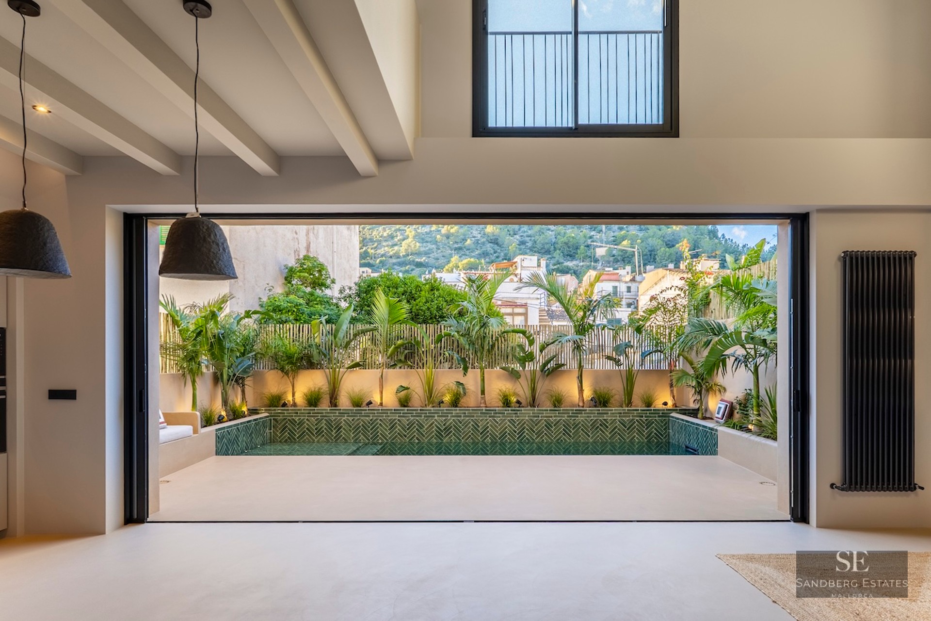 Vue intérieure par une grande baie vitrée vers une piscine privée entourée de plantes tropicales et carrelage vert.