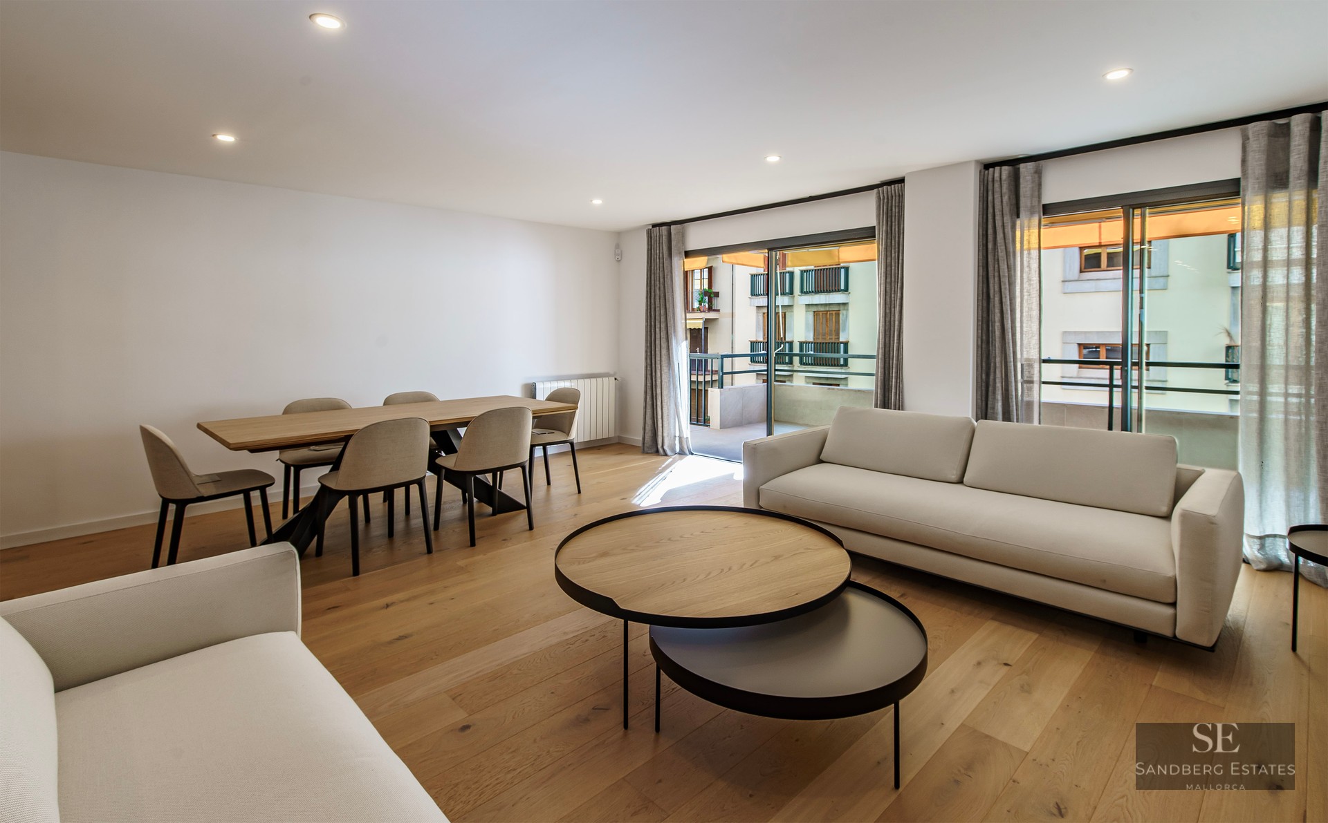 Salon moderne avec canapés blancs, tables basses en bois, coin repas et grandes baies vitrées ouvrant sur un balcon.