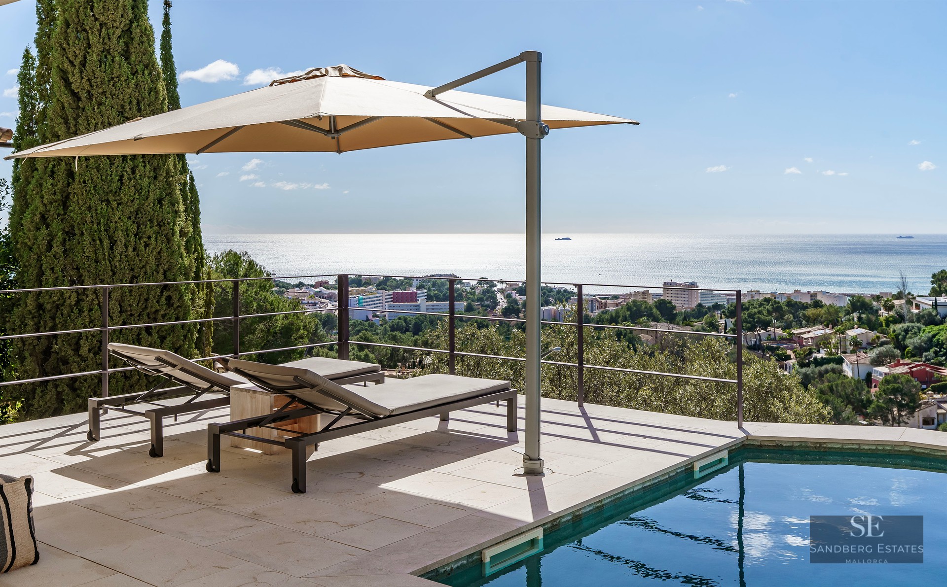 Modern pool terrace with two sun loungers under an umbrella, overlooking the sea and a coastal town.