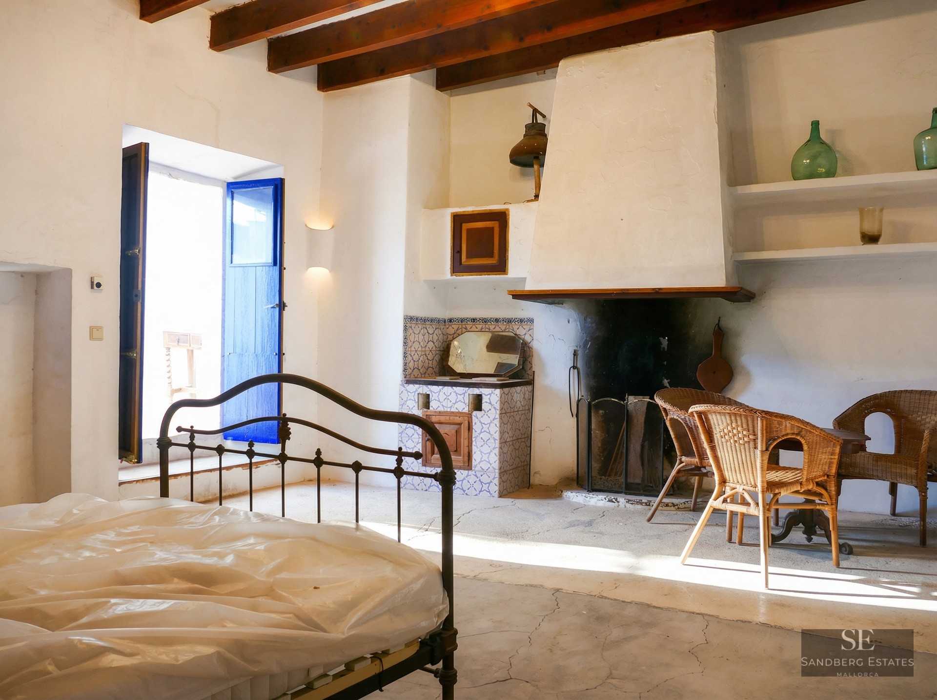 Bedroom with a wrought iron bed frame, a large white fireplace, blue wooden shutters, and wicker chairs under wooden beams.