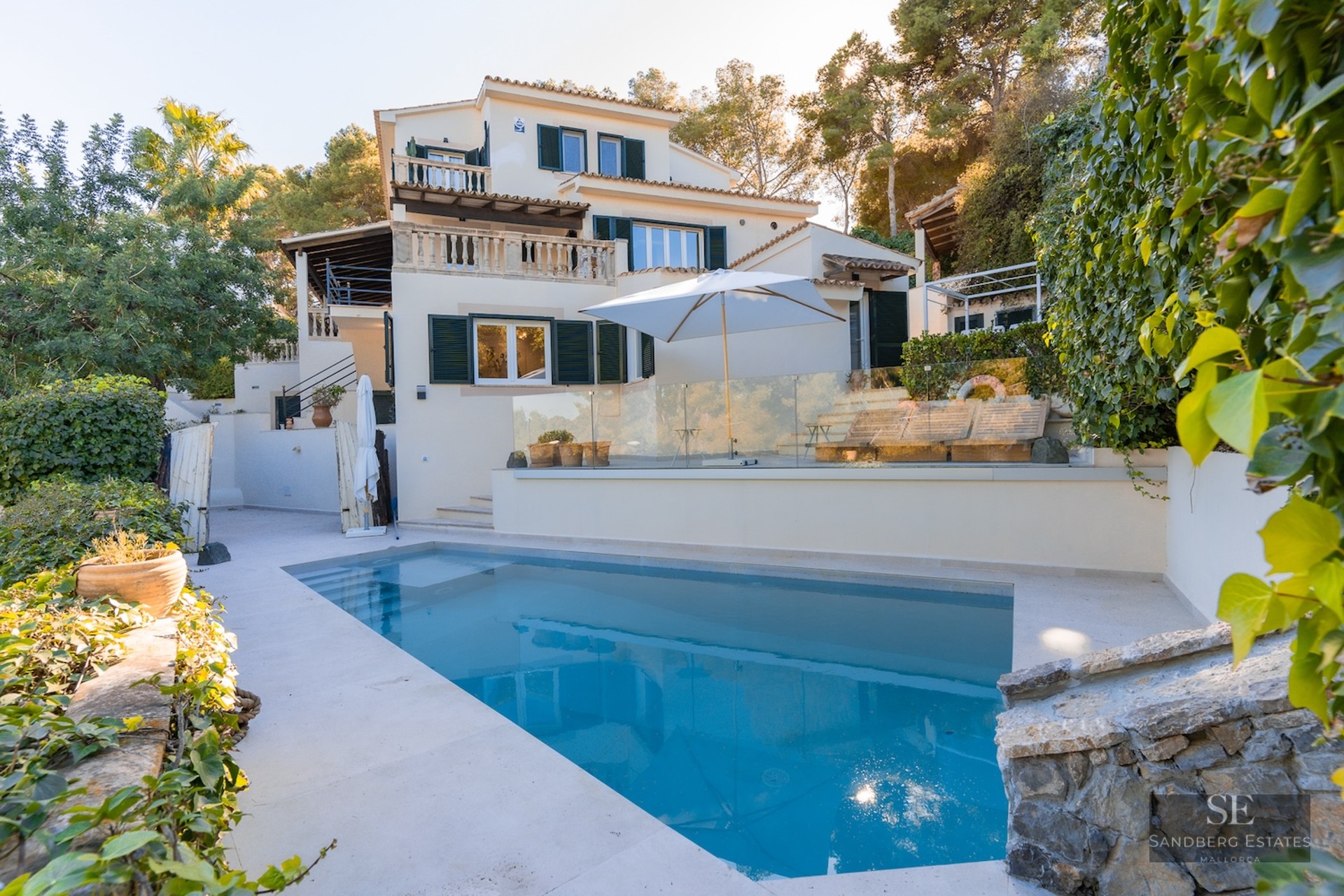 Rectangular swimming pool in the foreground of a multi-level white Mediterranean villa with green shutters and lush trees.
