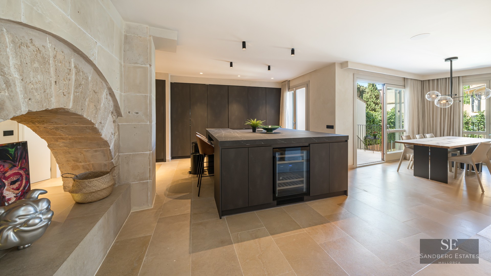 Modern kitchen with dark island and wine fridge next to a rustic stone arch and open-plan dining area.