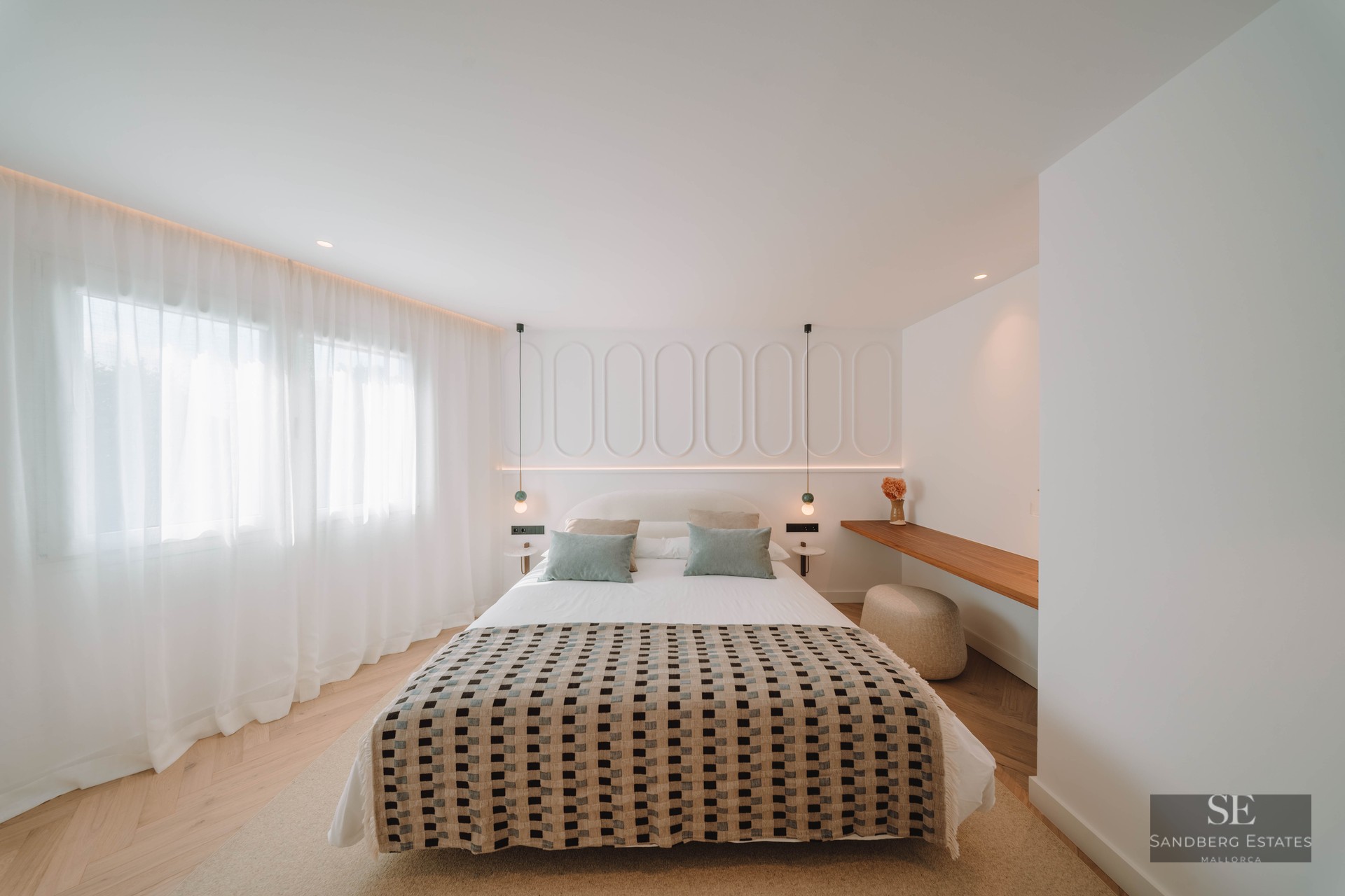 A bright, modern bedroom featuring a patterned bedspread, white paneled headboard wall, and herringbone wood flooring.