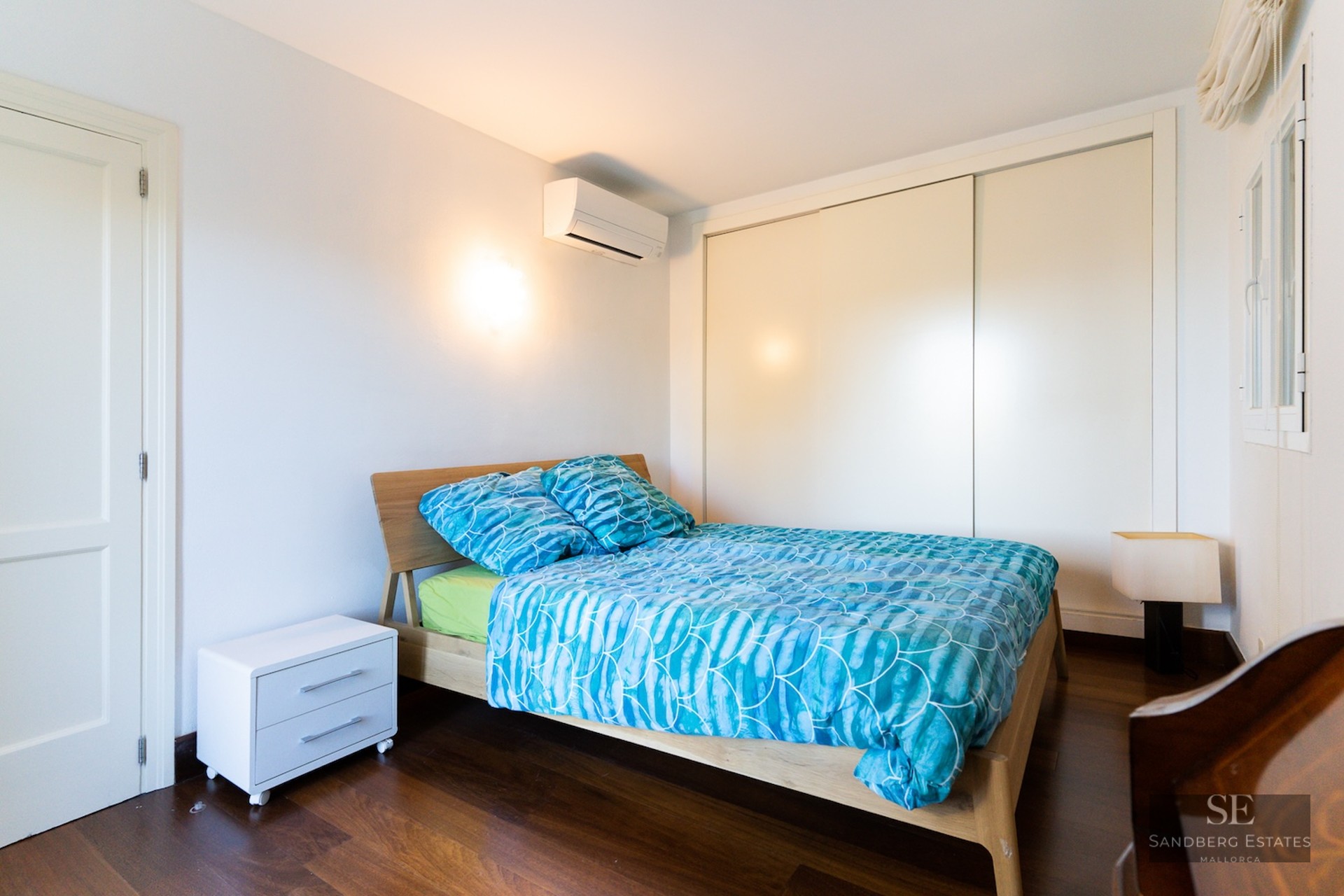 A modern bedroom with a wooden bed frame, blue patterned bedding, white walls, dark wood floors, and a built-in closet.