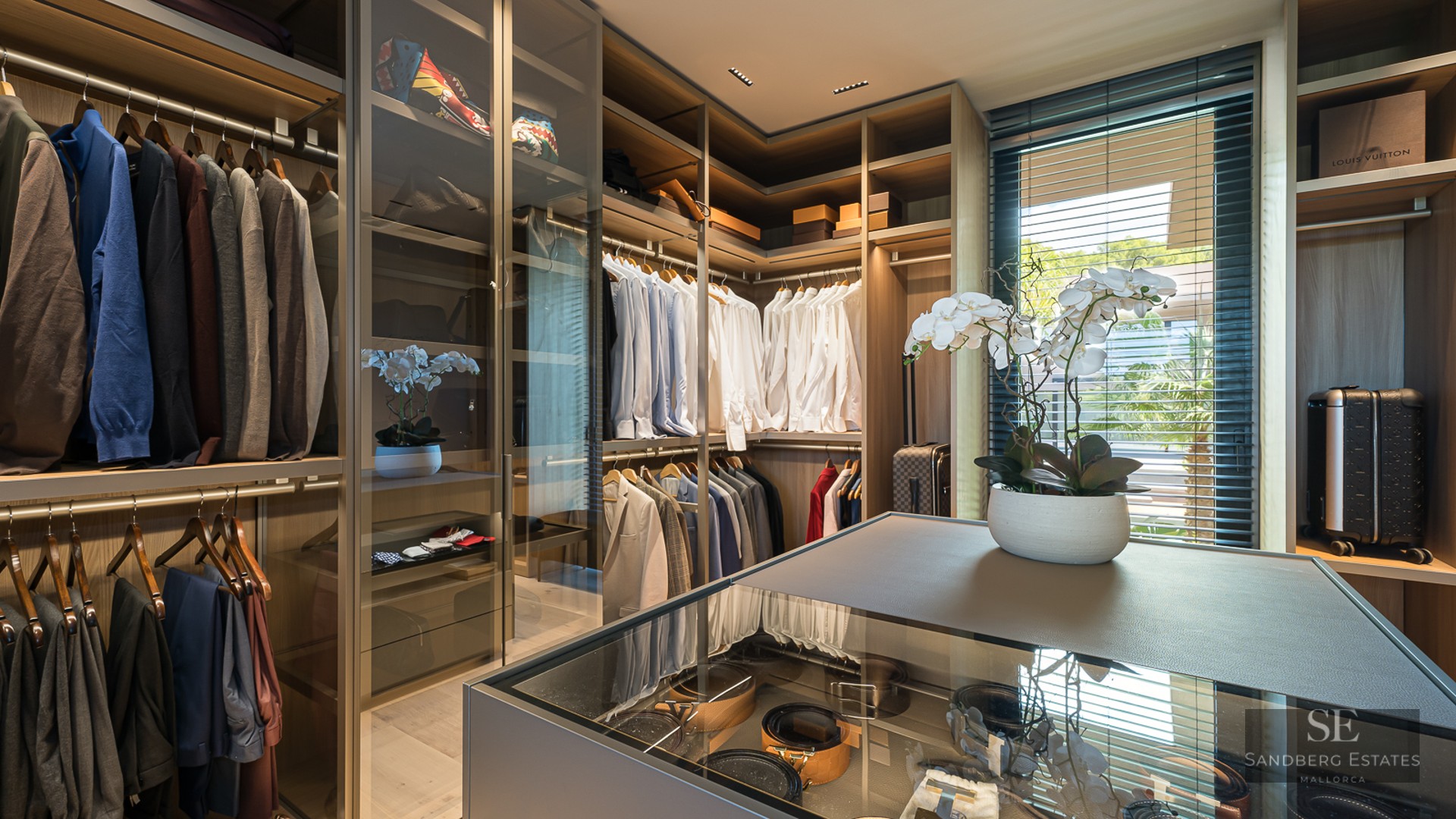 Spacious modern walk-in closet with wood shelving, glass display island, and designer clothes neatly organized.