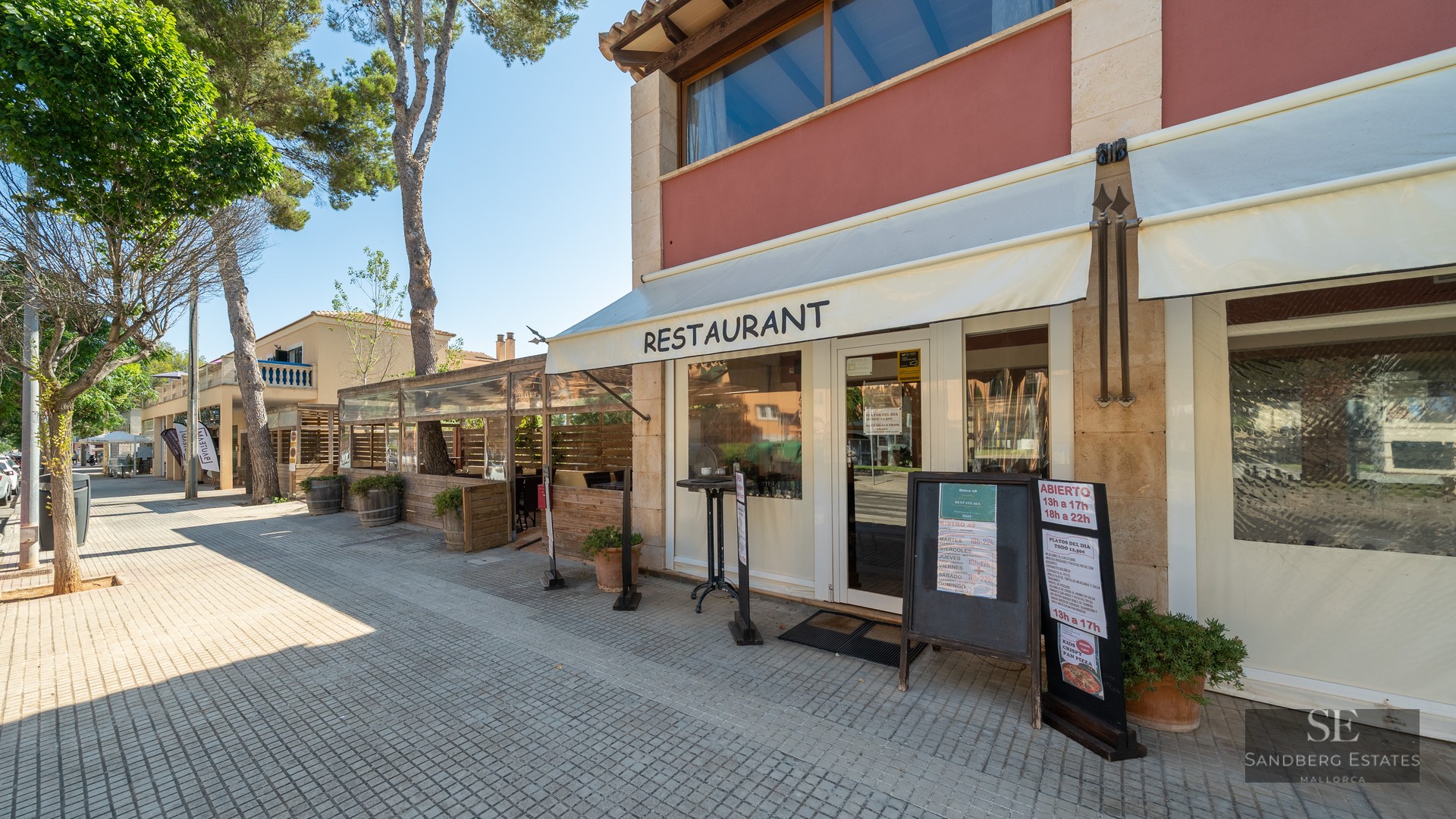 Exterior view of a restaurant with stone walls, white awnings, and a tree-lined sidewalk under a clear sky.