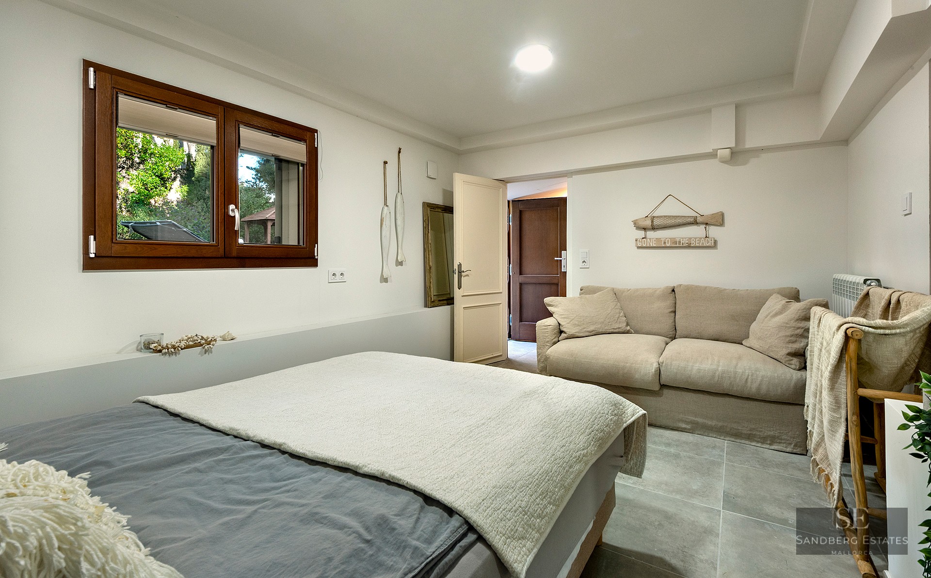 A bright bedroom featuring a grey bed, a beige sofa, a wooden window, and nautical-themed wall decor.