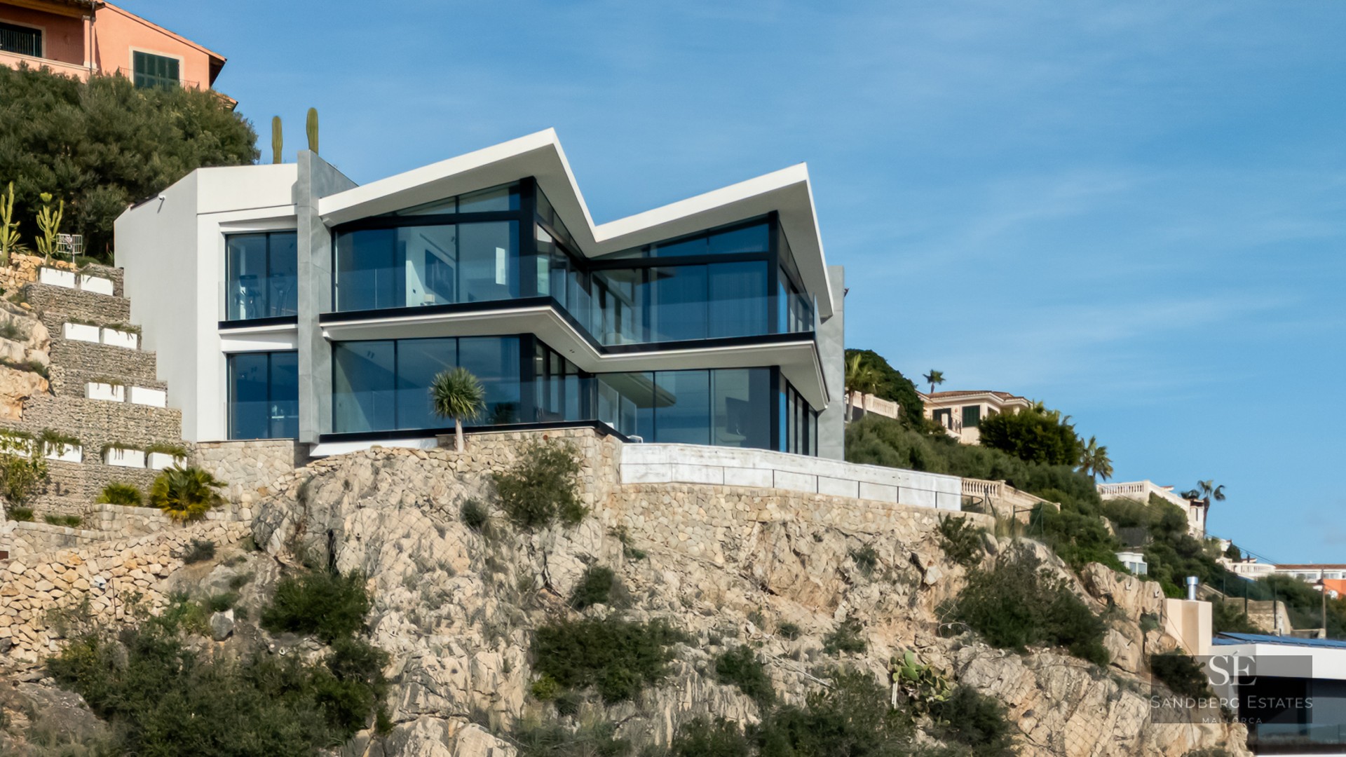 Modern two-story villa with sharp angular roofs and large glass walls built into a rocky hillside under a blue sky.