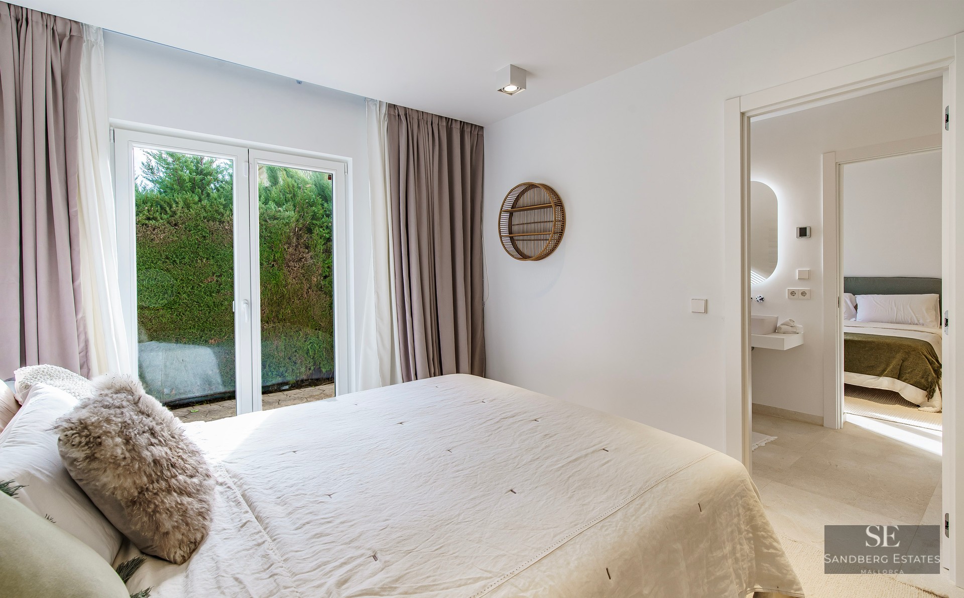 Bright master bedroom with white linens, glass doors to a garden, and a view into an ensuite bathroom.