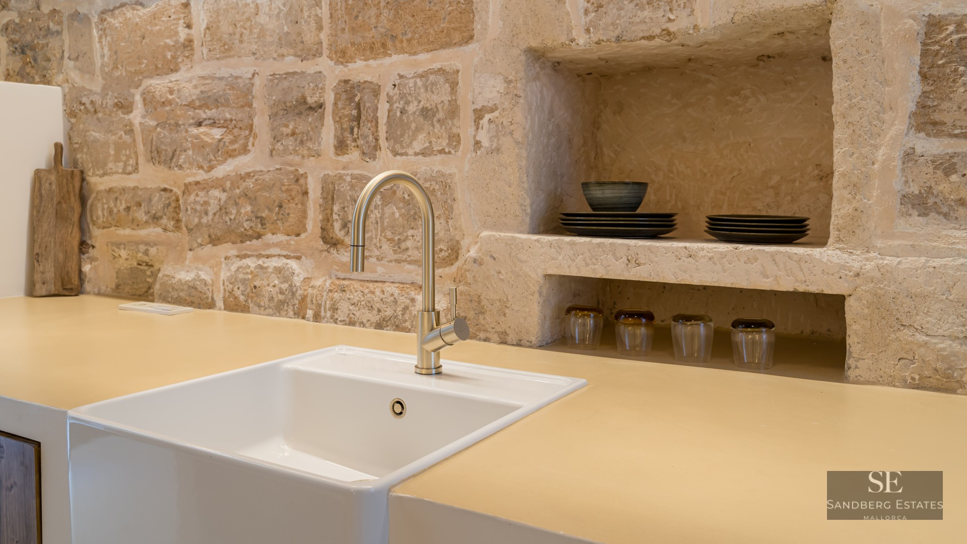 Modern gold faucet and white farmhouse sink against a rustic exposed stone wall with built-in shelving.