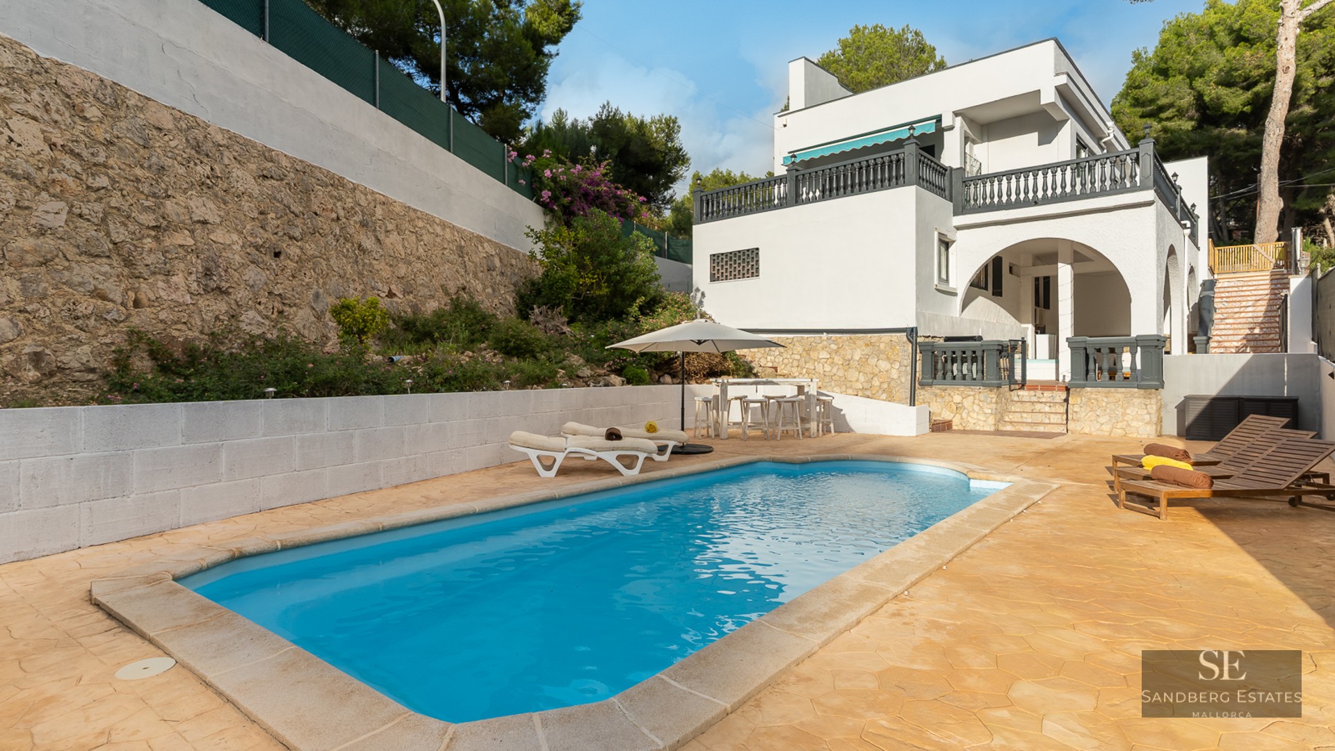 Piscine rectangulaire à l'eau turquoise entourée d'une terrasse en pierre, de chaises longues et d'une villa blanche.