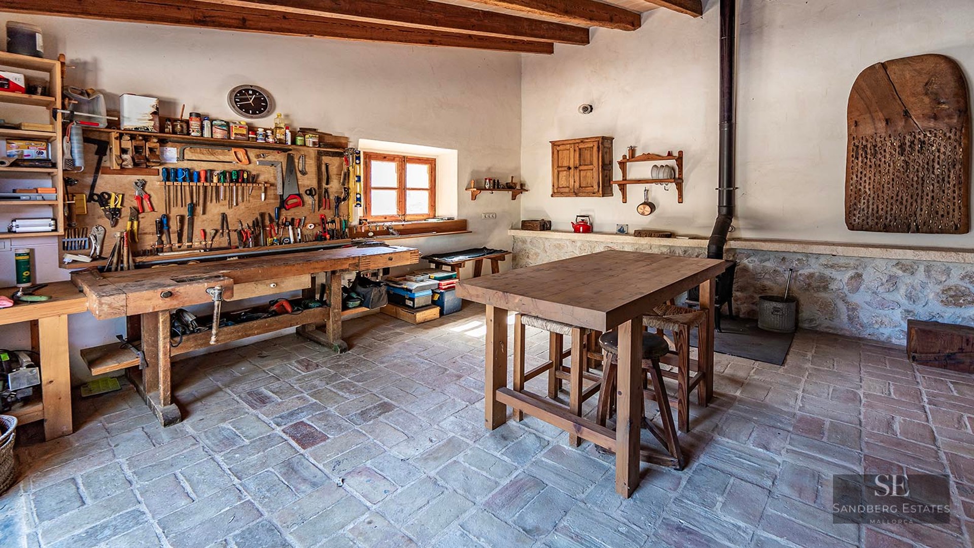 A traditional workshop with a large wooden workbench, tools on the wall, and rustic stone tile floors.